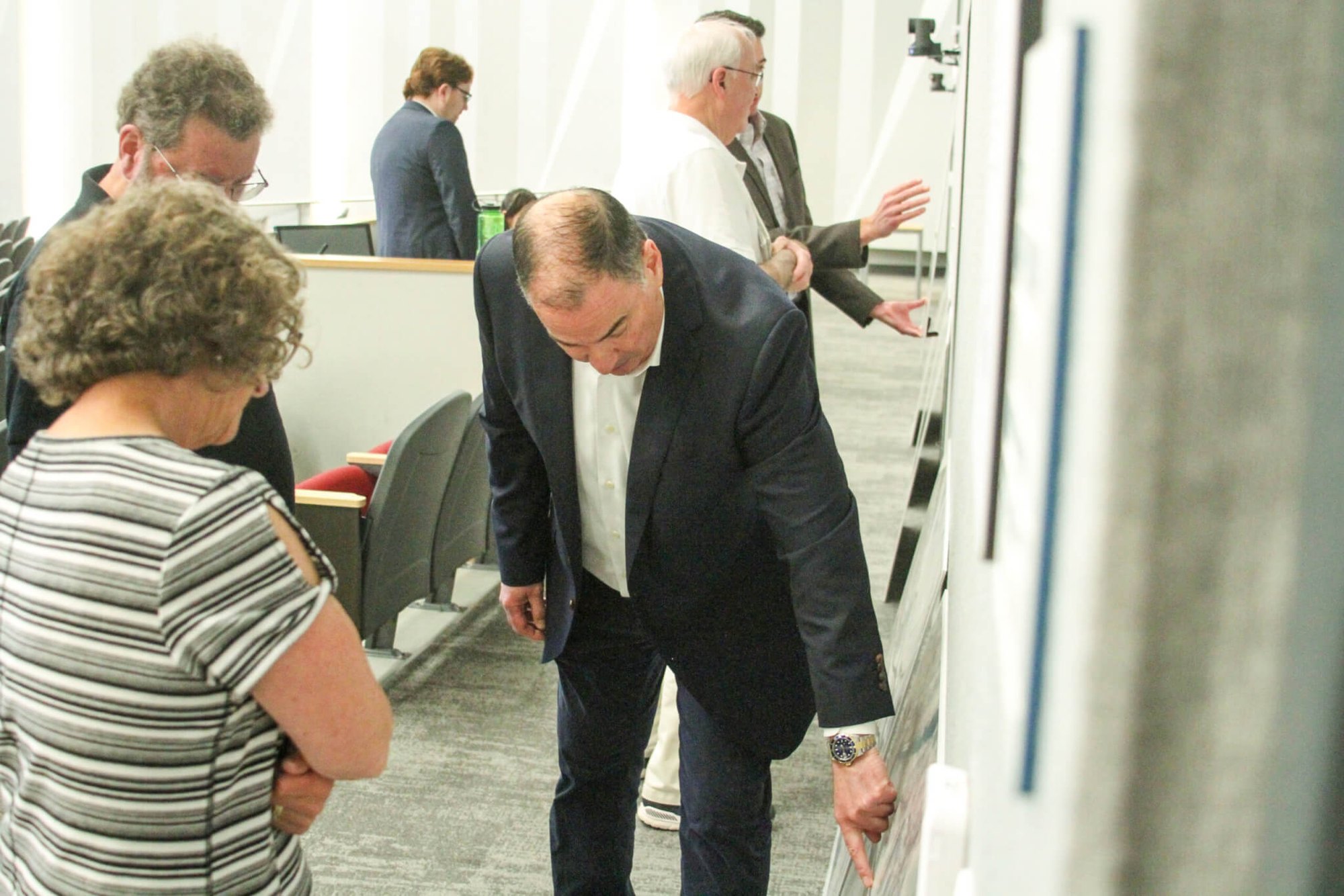 A man in a dark suit points at a large design poster while a woman in a striped top observes at a public meeting.