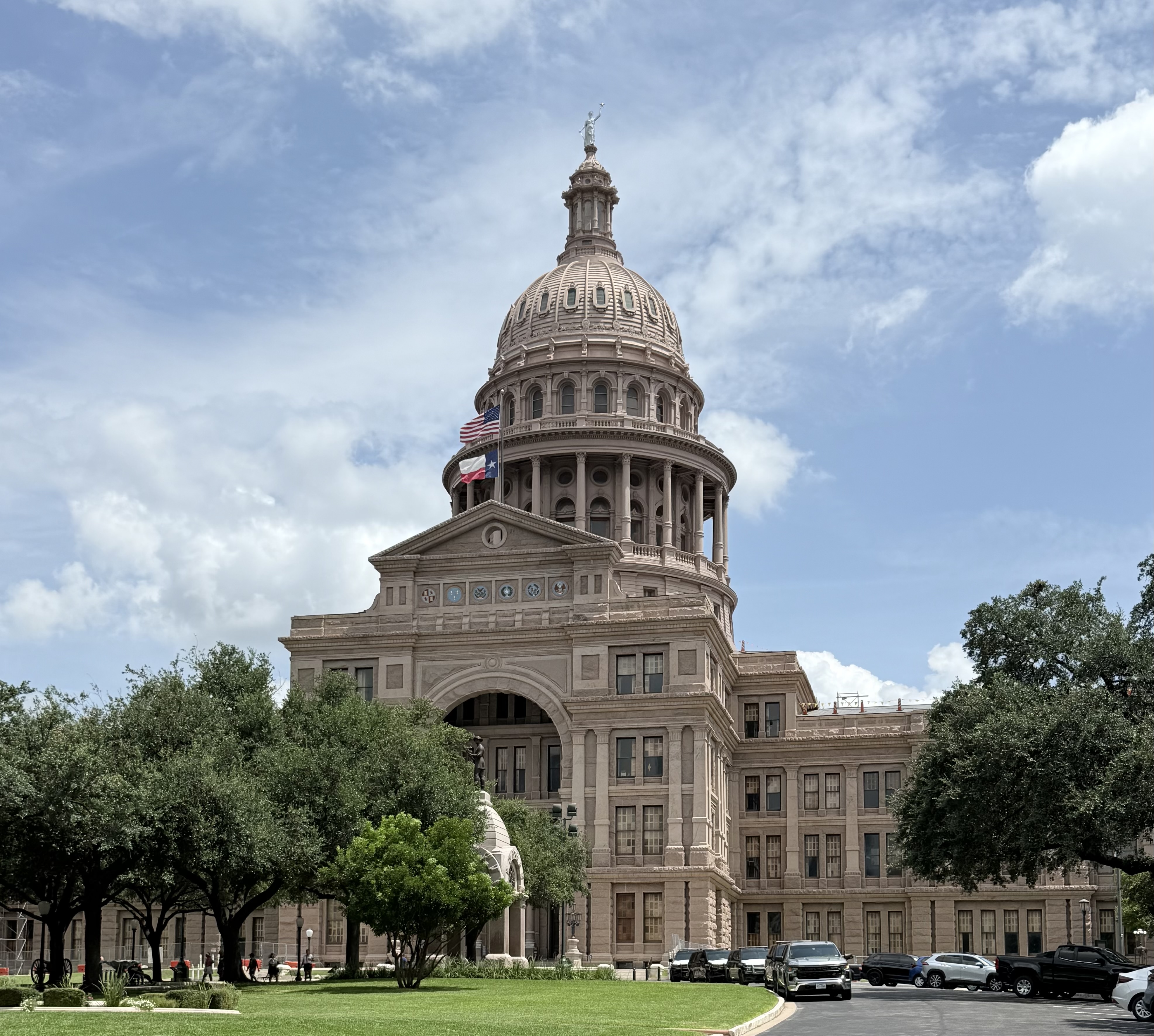 Texas State Capitol building with dome and two flags, framed by trees.