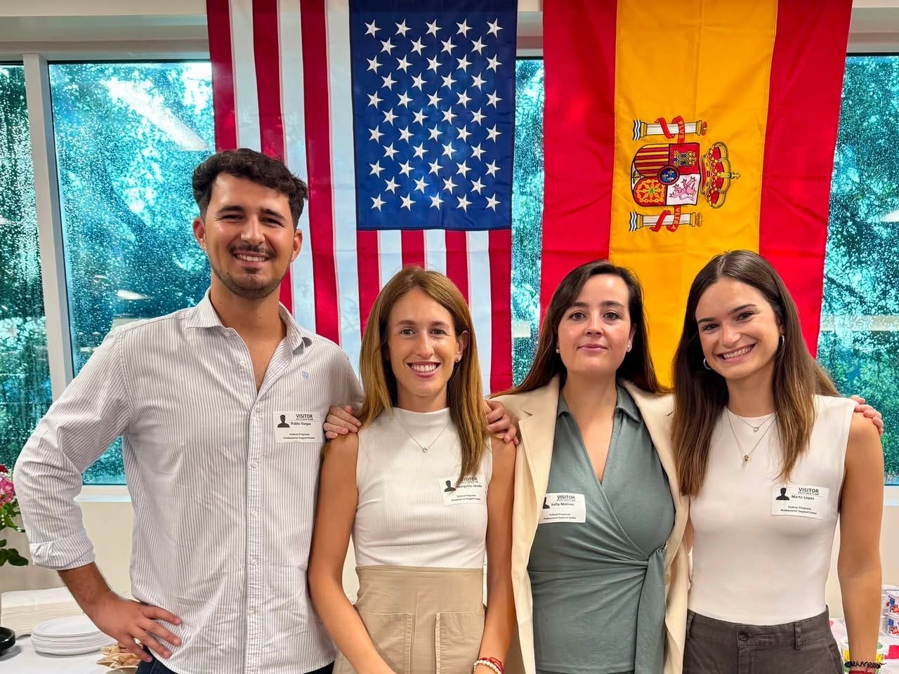Four smiling adults wearing name badges stand in front of an American flag and a Spanish flag indoors.