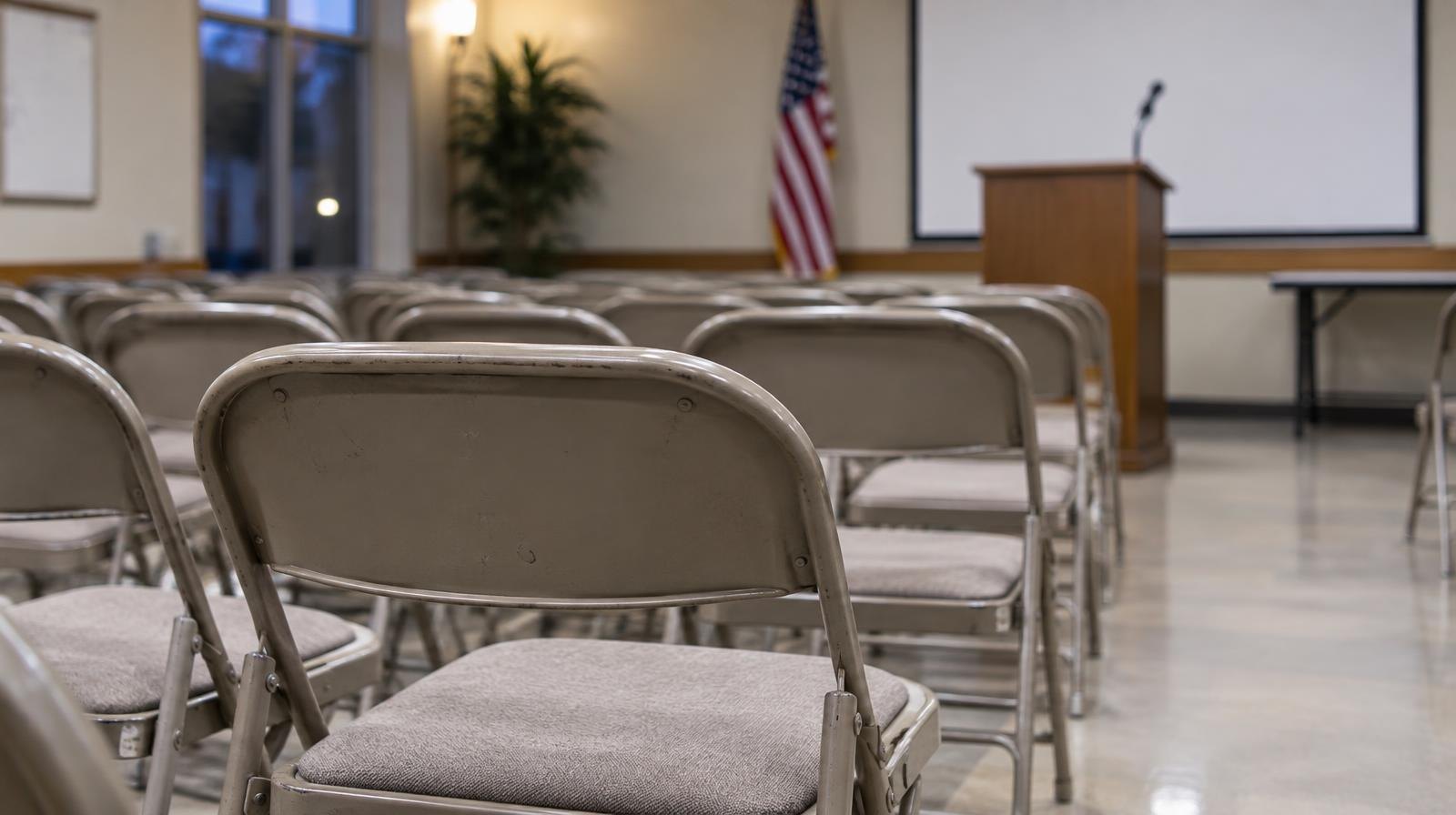 Rows of empty folding chairs face a podium in a meeting room.