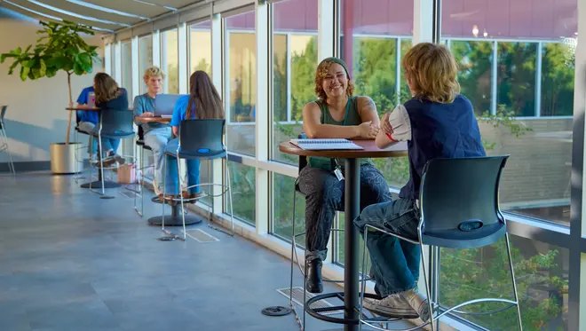 Two students sit at a tall table and talk in a bright, windowed college student lounge.