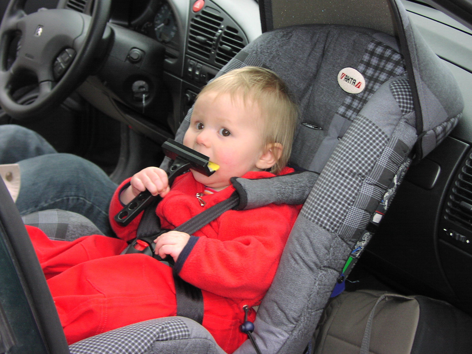 An infant in a red outfit buckled into a rear-facing car seat inside a vehicle.