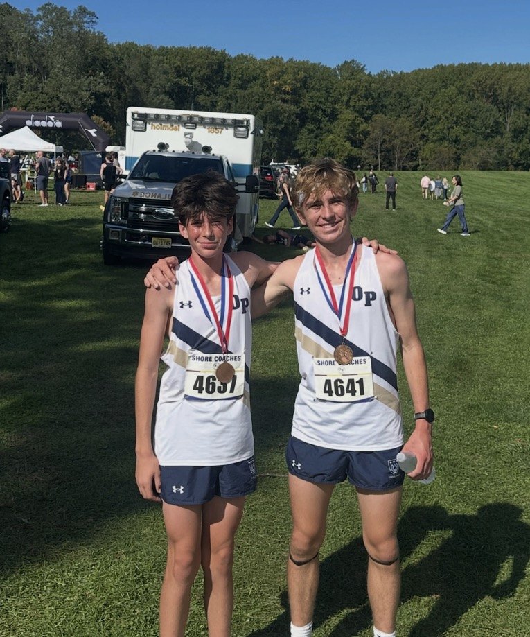 Two runners in white uniforms with race bibs and medals pose on a grass field.