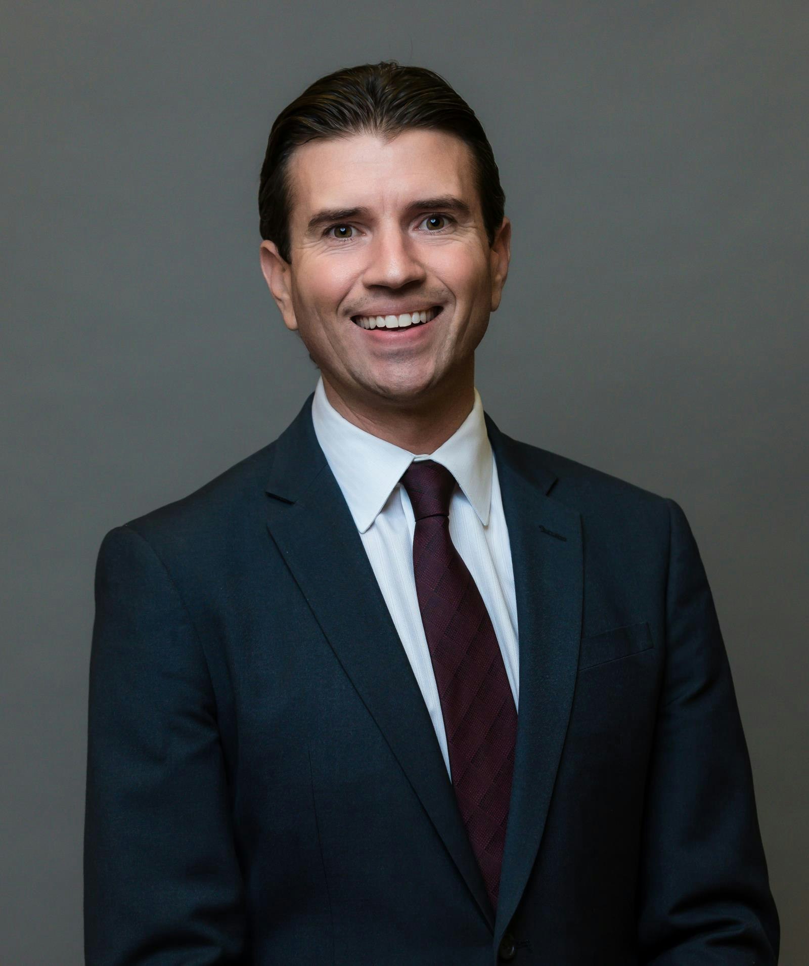 Man in a dark suit and burgundy tie smiling in a professional headshot against a gray background.