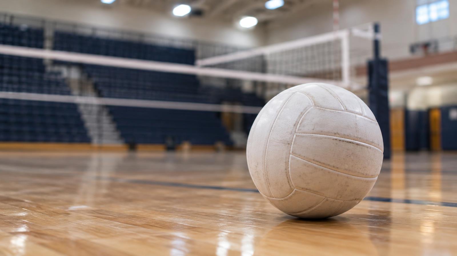 A white volleyball on a hardwood gymnasium floor near the net.