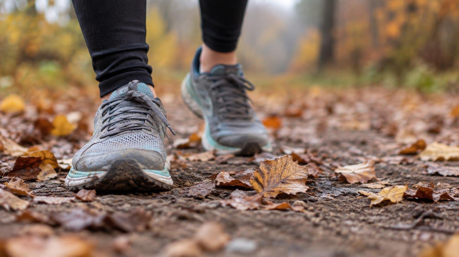 A pair of running shoes on a dirt trail surrounded by fallen autumn leaves.