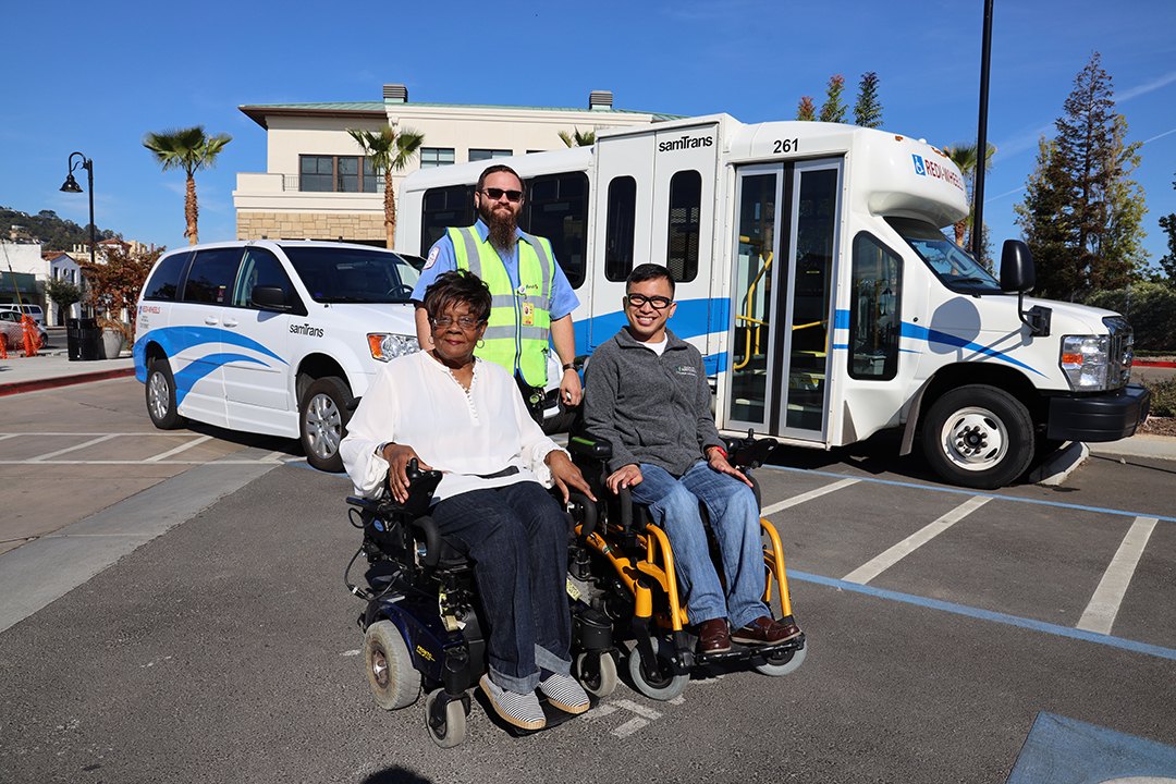 Two wheelchair users and a worker in a safety vest stand in front of accessible transit vans.