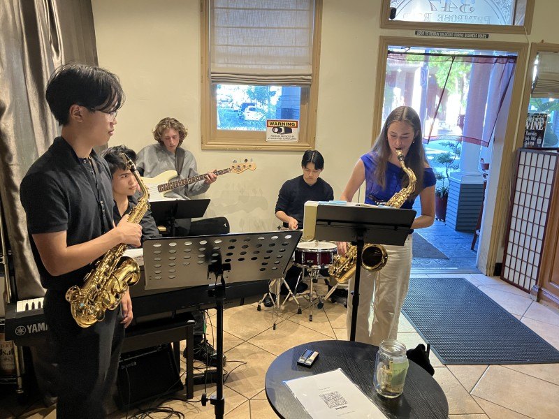Young musicians play saxophones and bass inside a casual restaurant, reading from music stands.