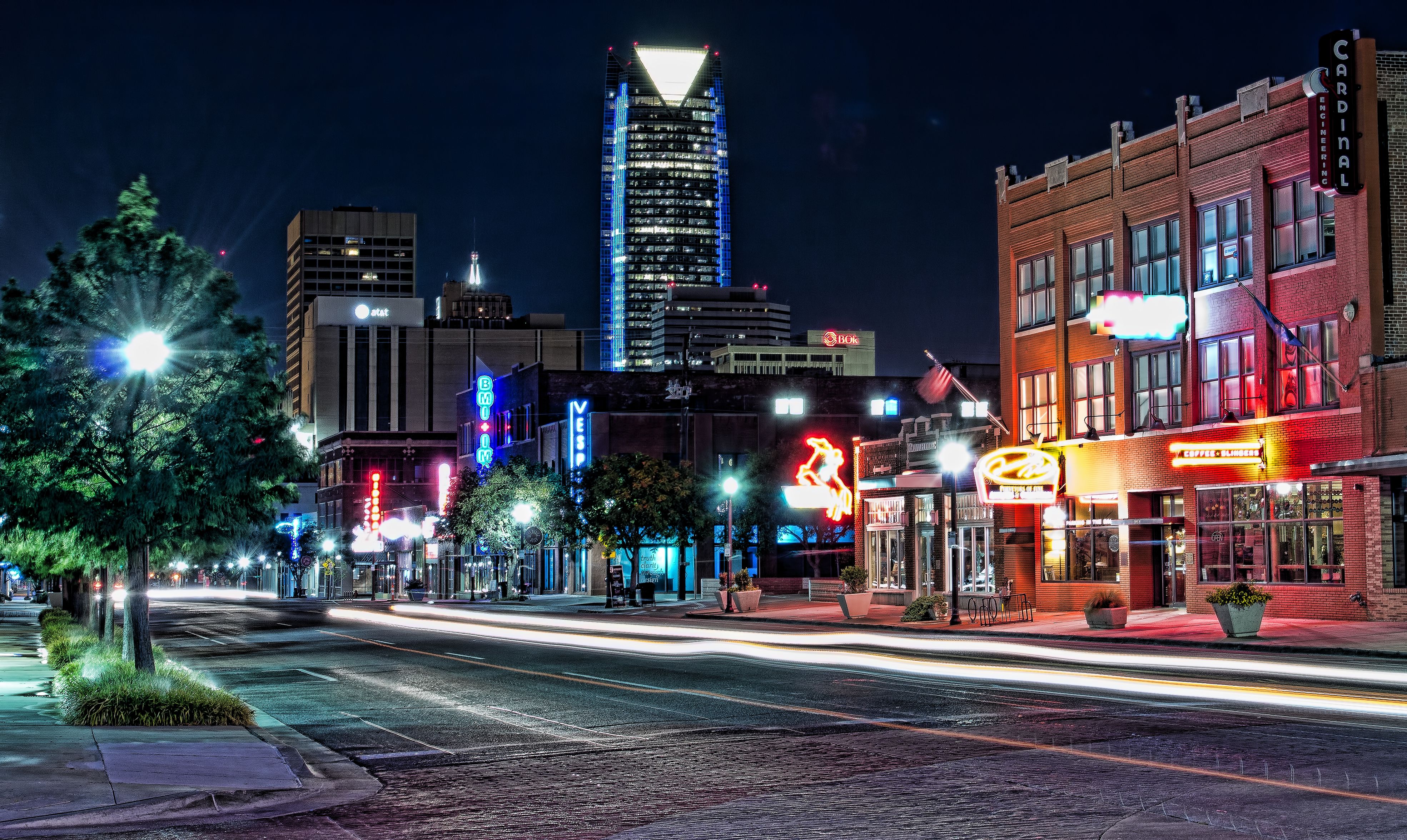 Nighttime street in downtown Oklahoma City with neon signs, brick storefronts, and a lit skyscraper.