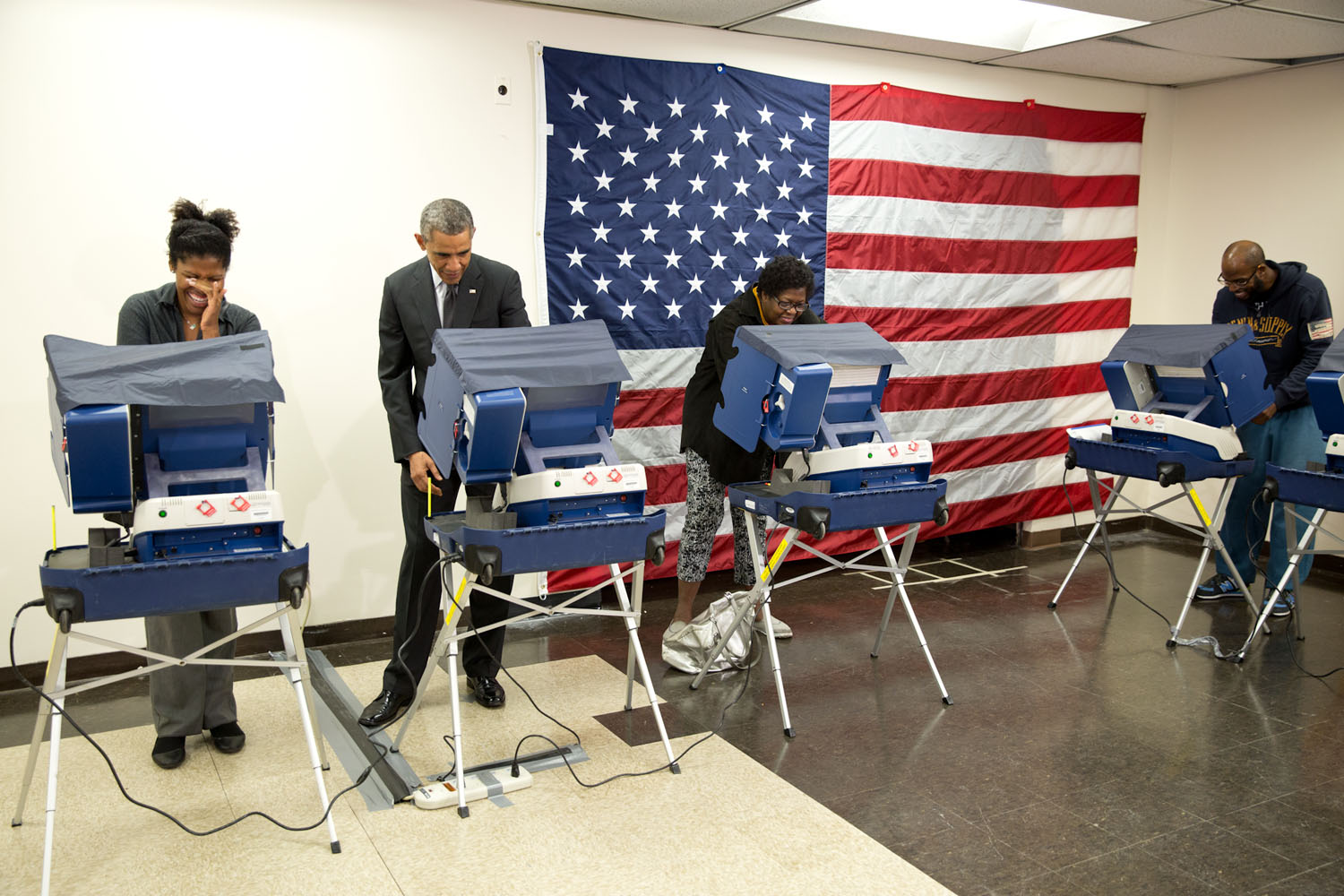 Four people using electronic voting machines at an indoor polling location with an American flag on the wall.