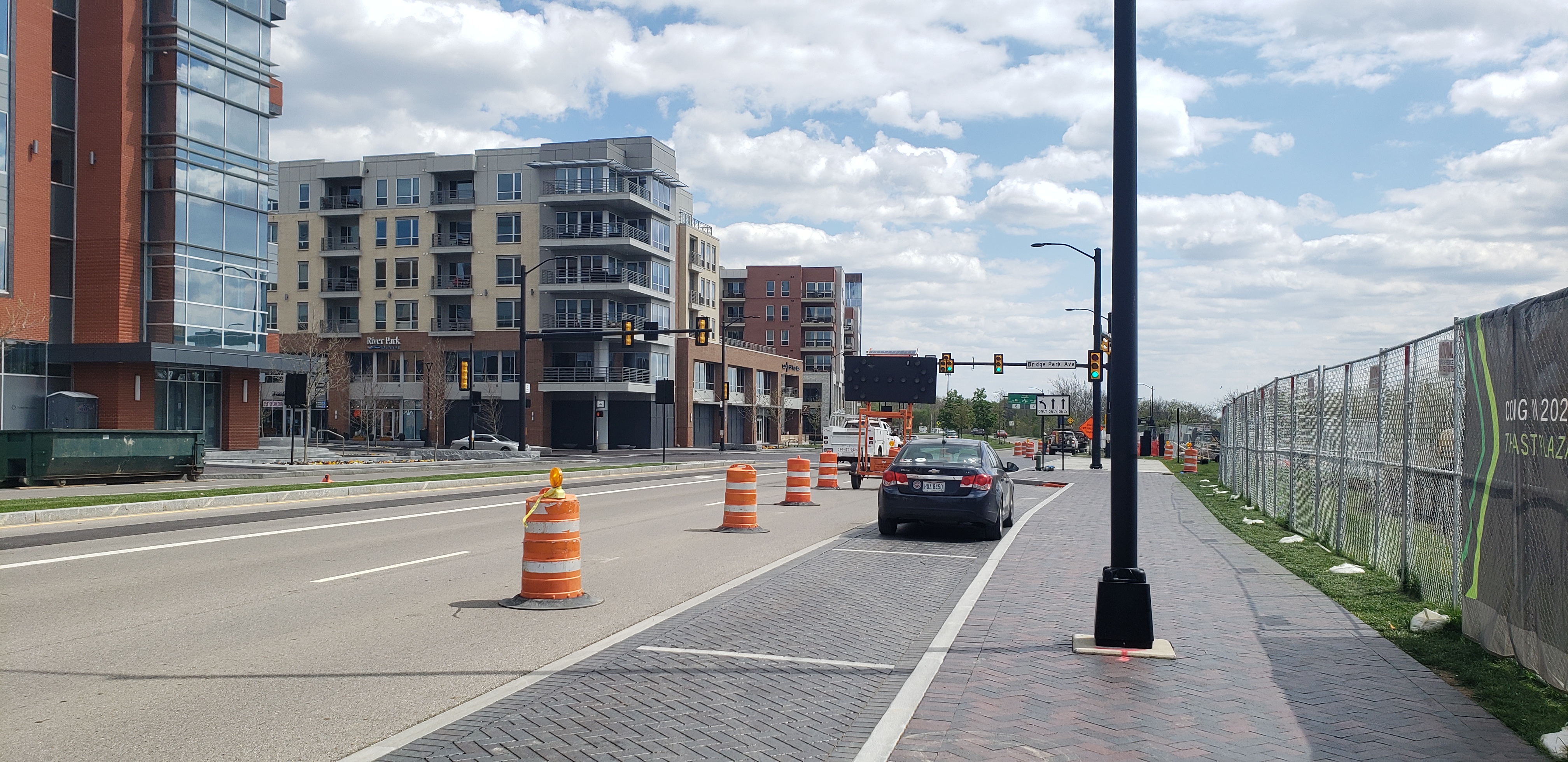 Urban road with orange construction barrels, a car in traffic, and modern buildings behind.