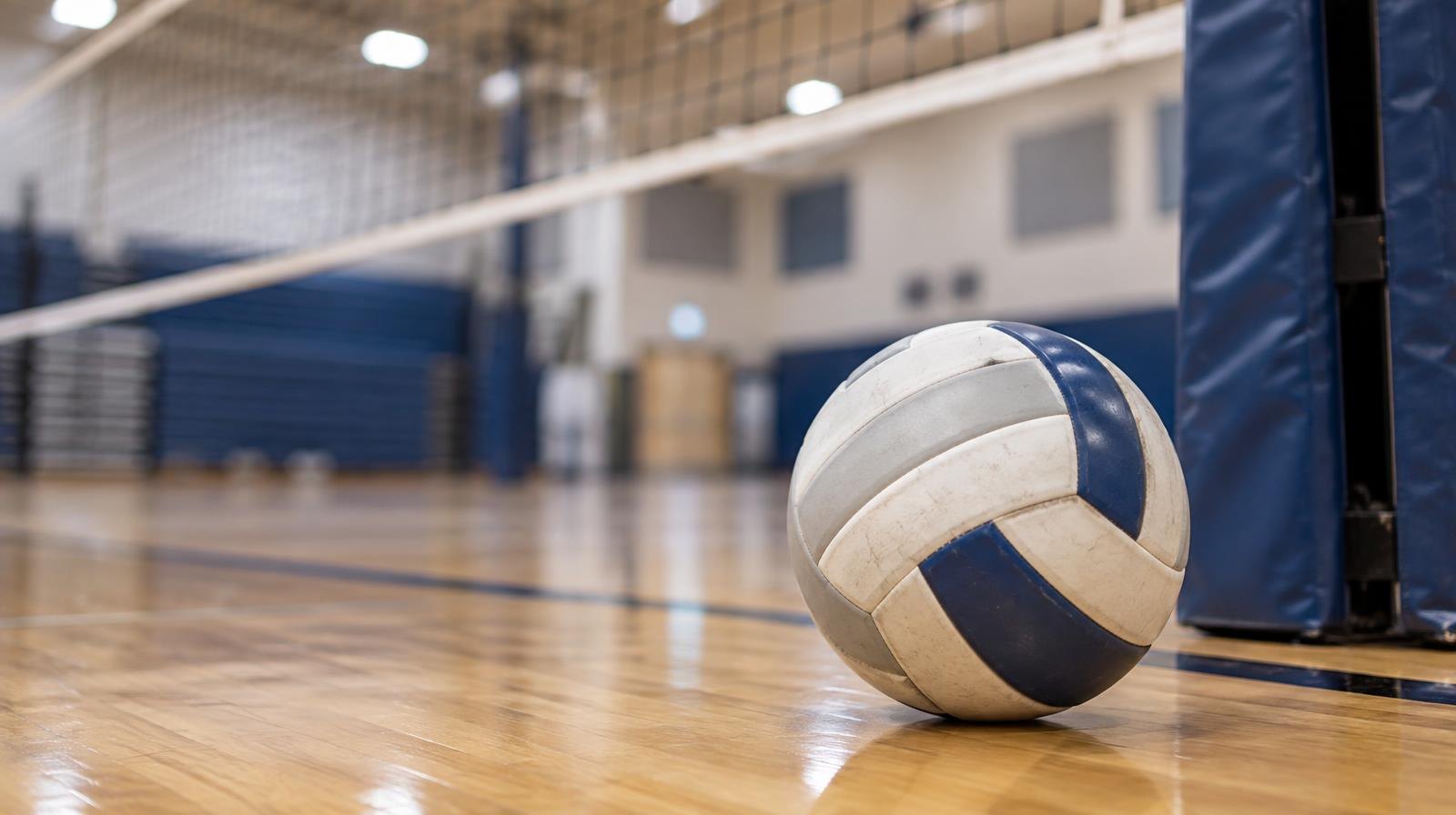 A volleyball resting on a hardwood gym floor near a net post under bright overhead lights.