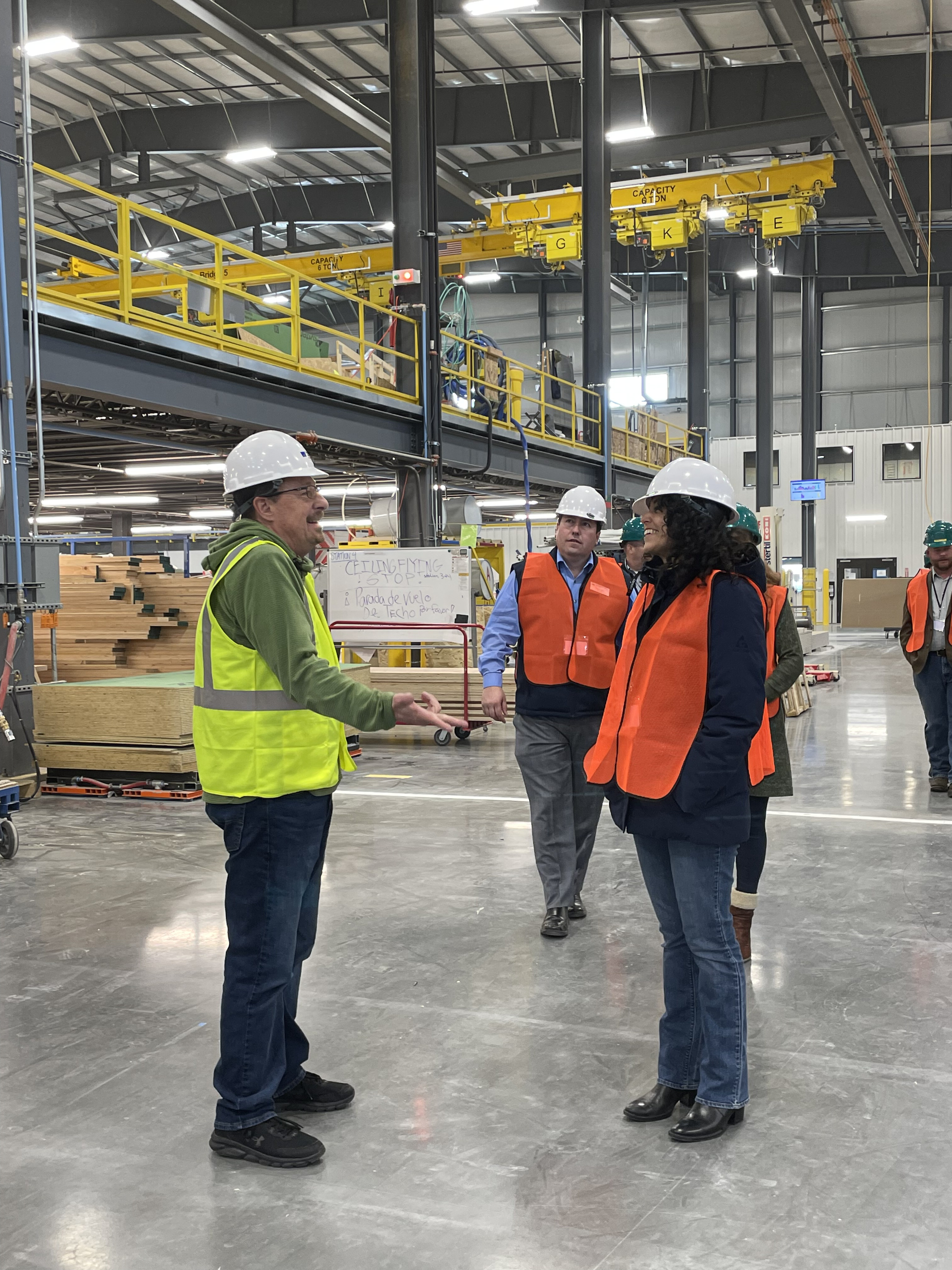 Three workers in hard hats and safety vests stand on a large factory floor with overhead cranes.