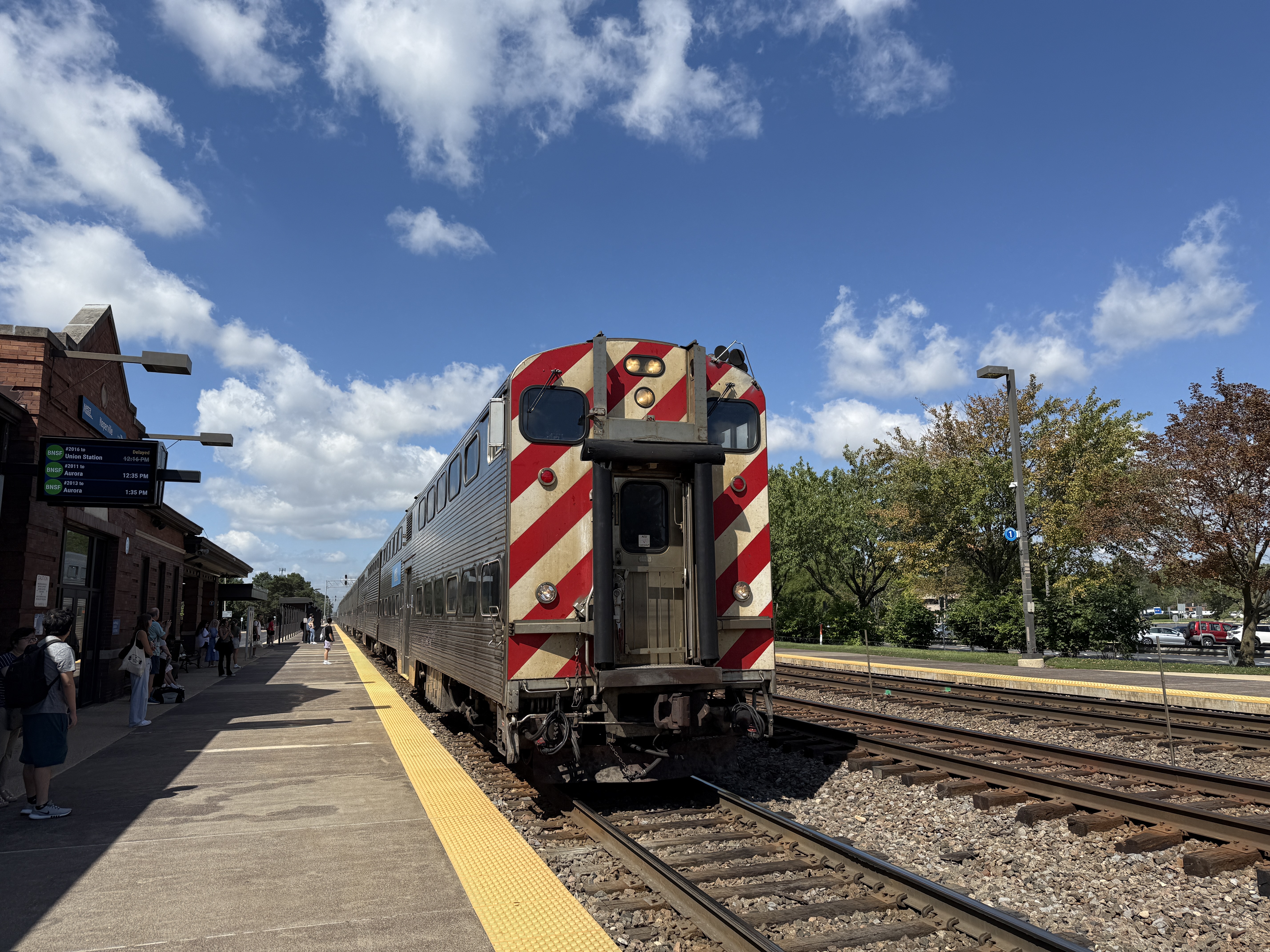 A Metra train pulls into a station platform beside a brick station building with commuters waiting.