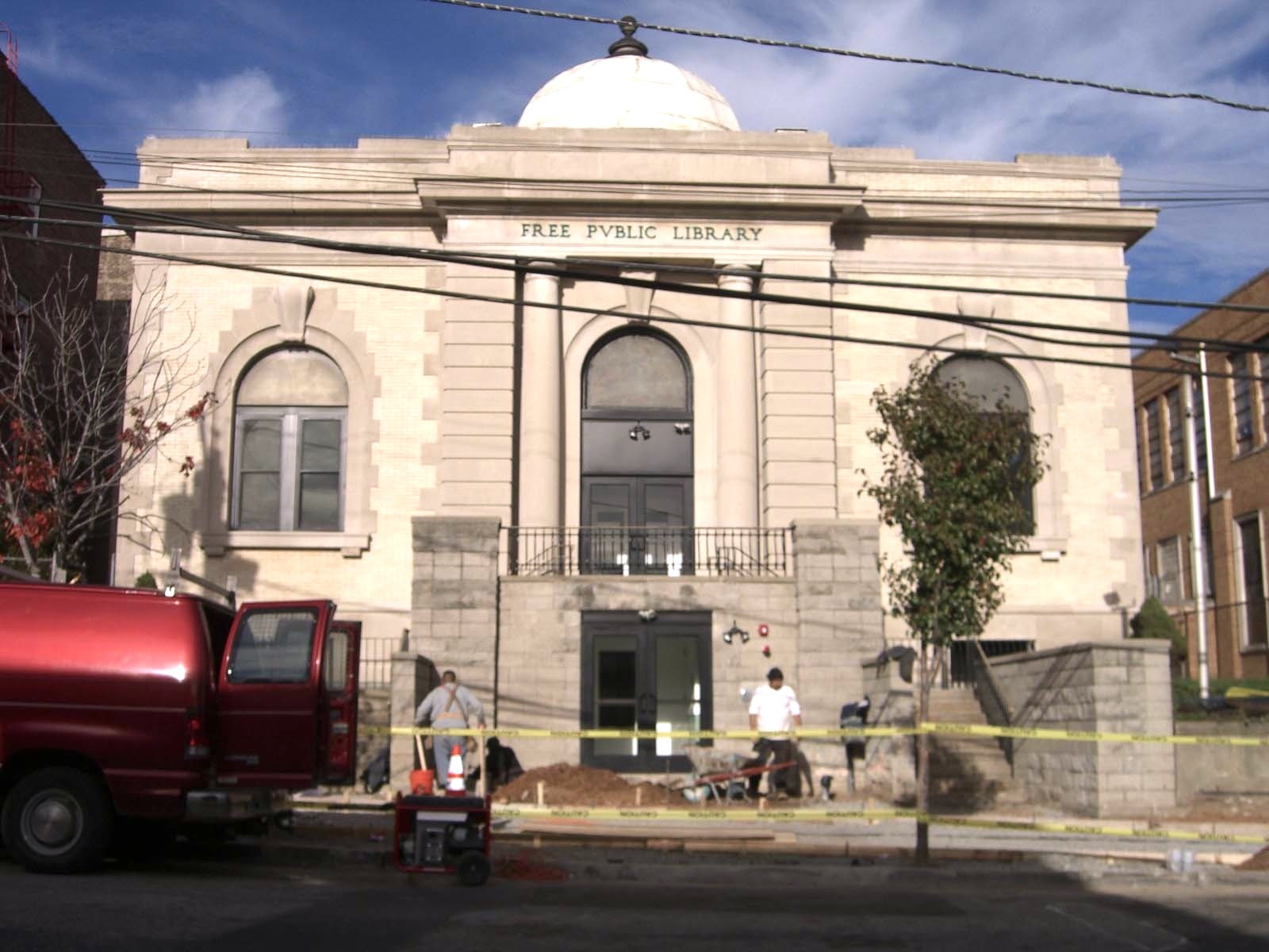 Construction workers and equipment in front of a neoclassical public library building with yellow caution tape.