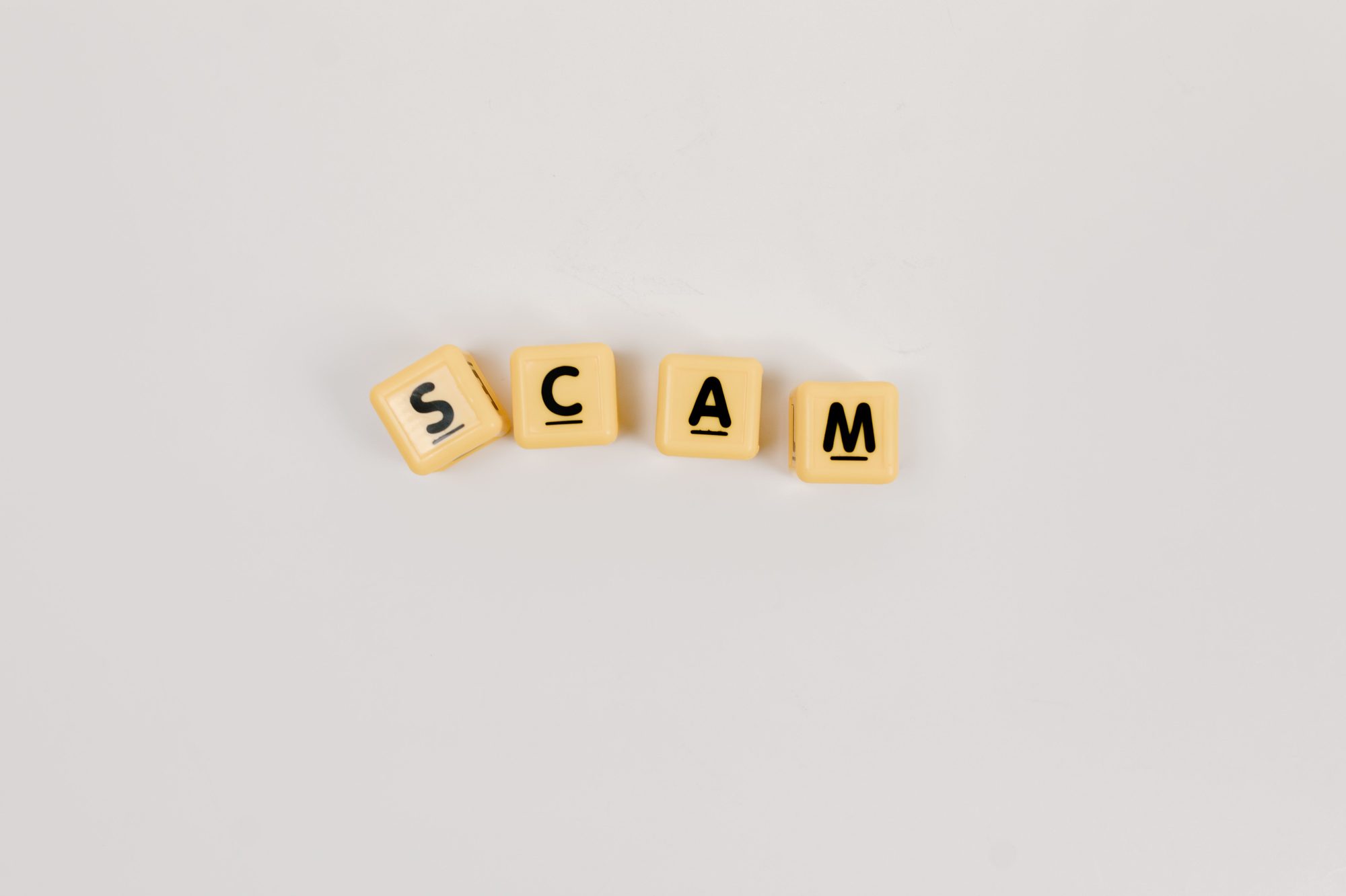 Four Scrabble tiles forming the word 'SCAM' on a neutral background, highlighting concepts like deceit.