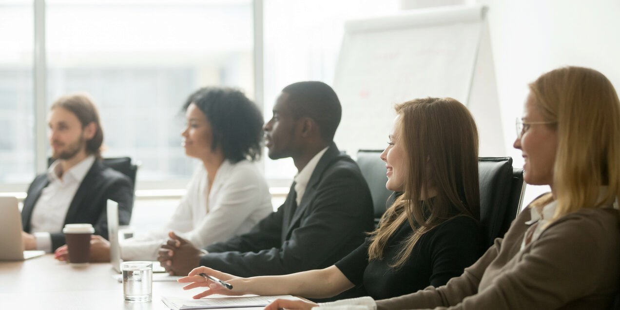 Five professionally dressed people of different backgrounds seated at a conference table in a meeting room.