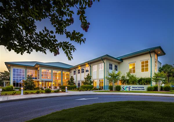 Modern two-story college building with teal metal roof and glass facade lit at dusk.