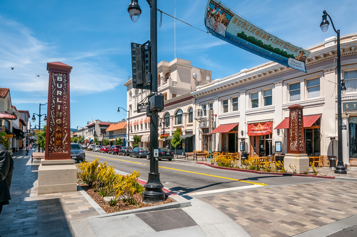 Sunny commercial street with a decorative pillar sign reading 'Burlingame' and storefronts with red awnings.
