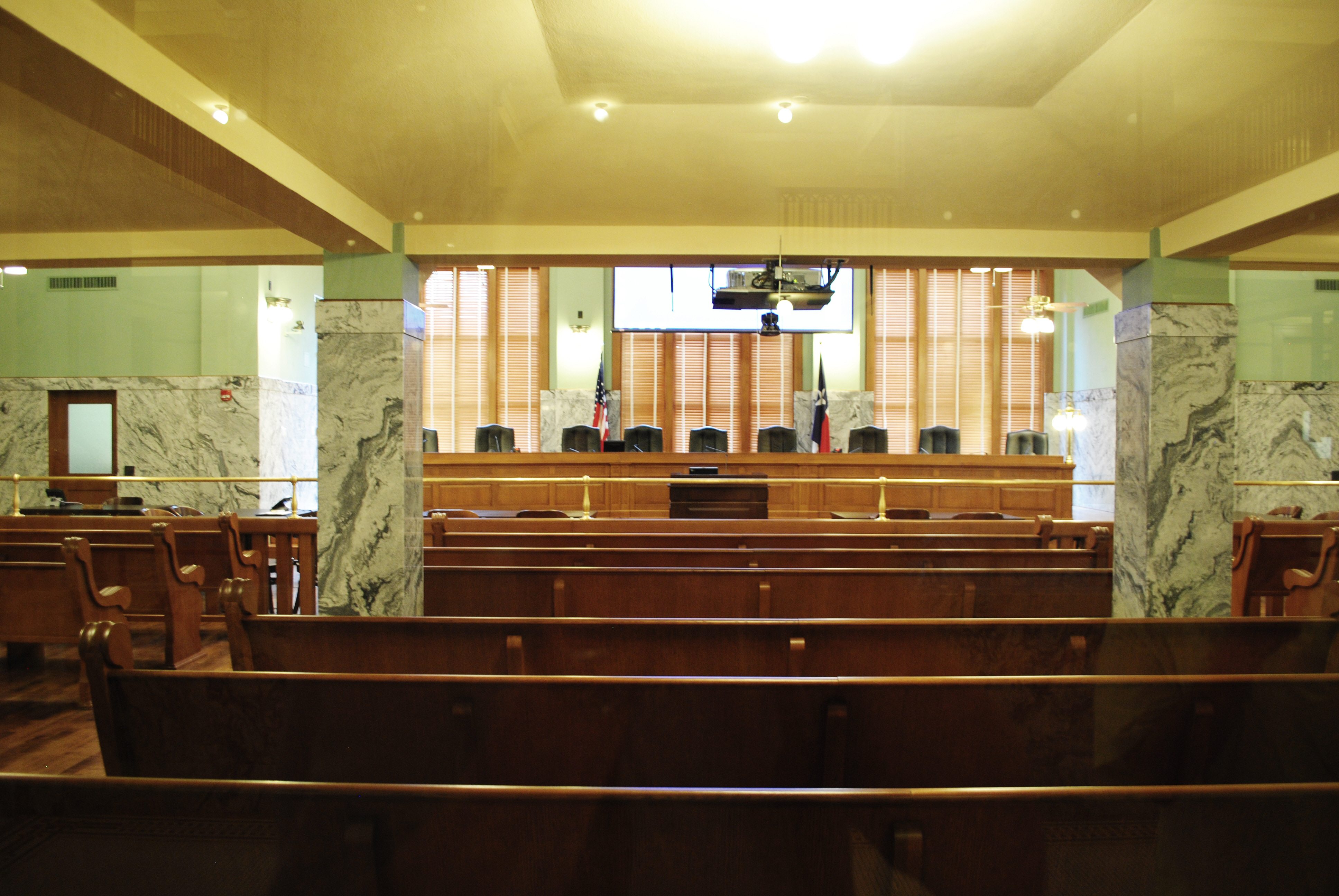 Empty government chamber with raised bench, wooden pews, marble columns, and two flags.