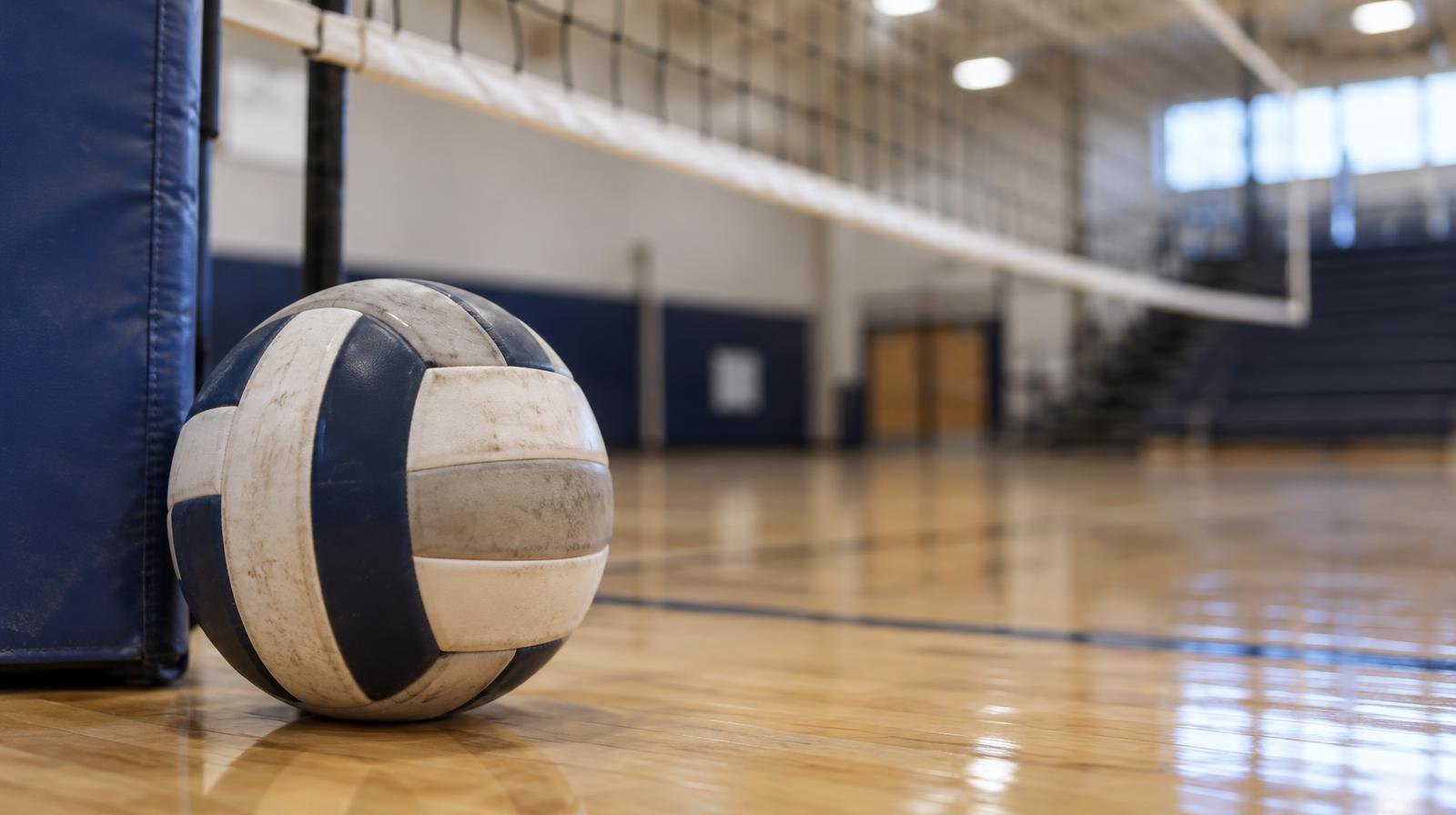 A volleyball resting on a hardwood gymnasium floor near the net post.