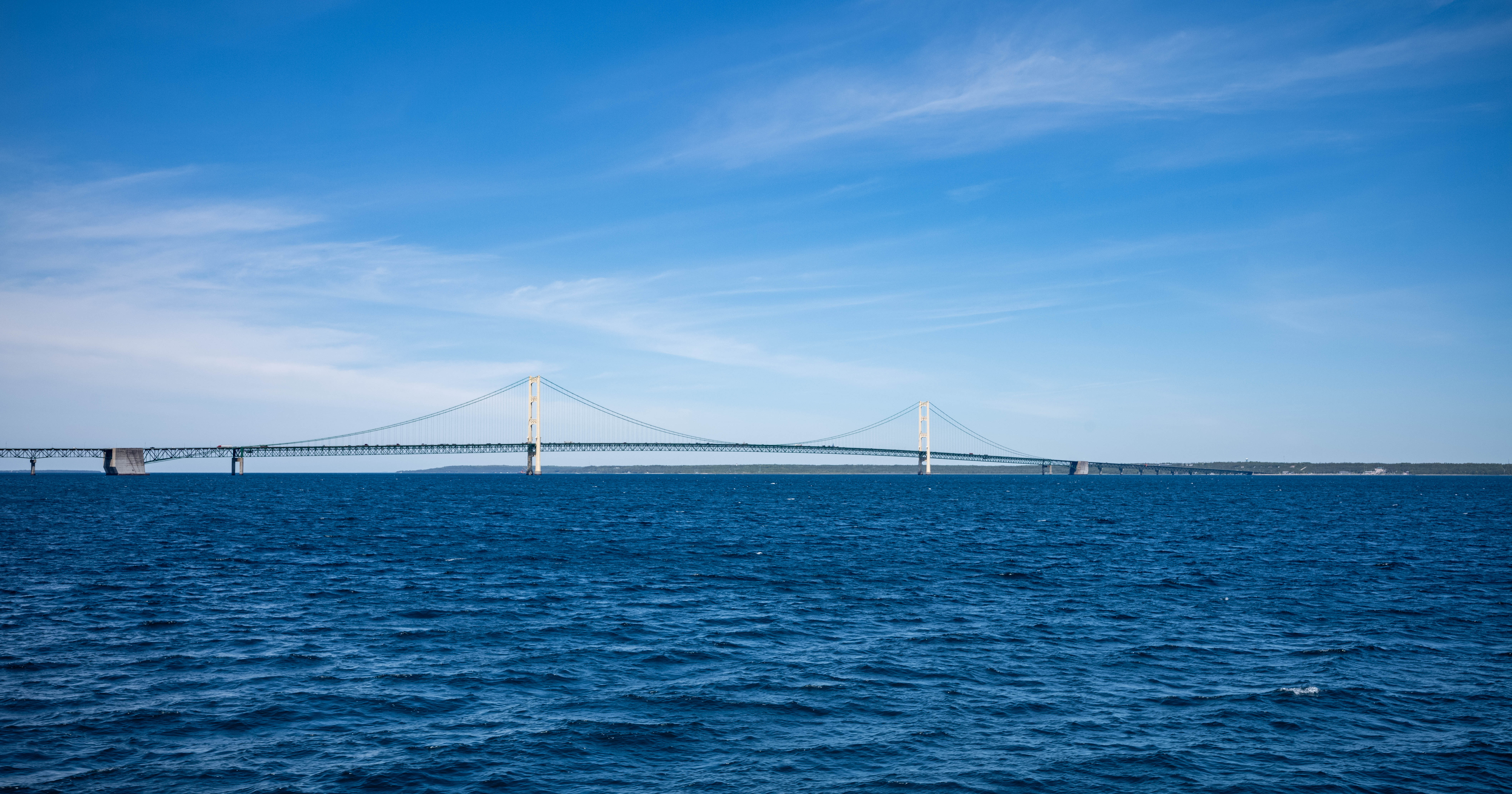 Long suspension bridge spanning blue water under a partly cloudy sky, viewed from water level.