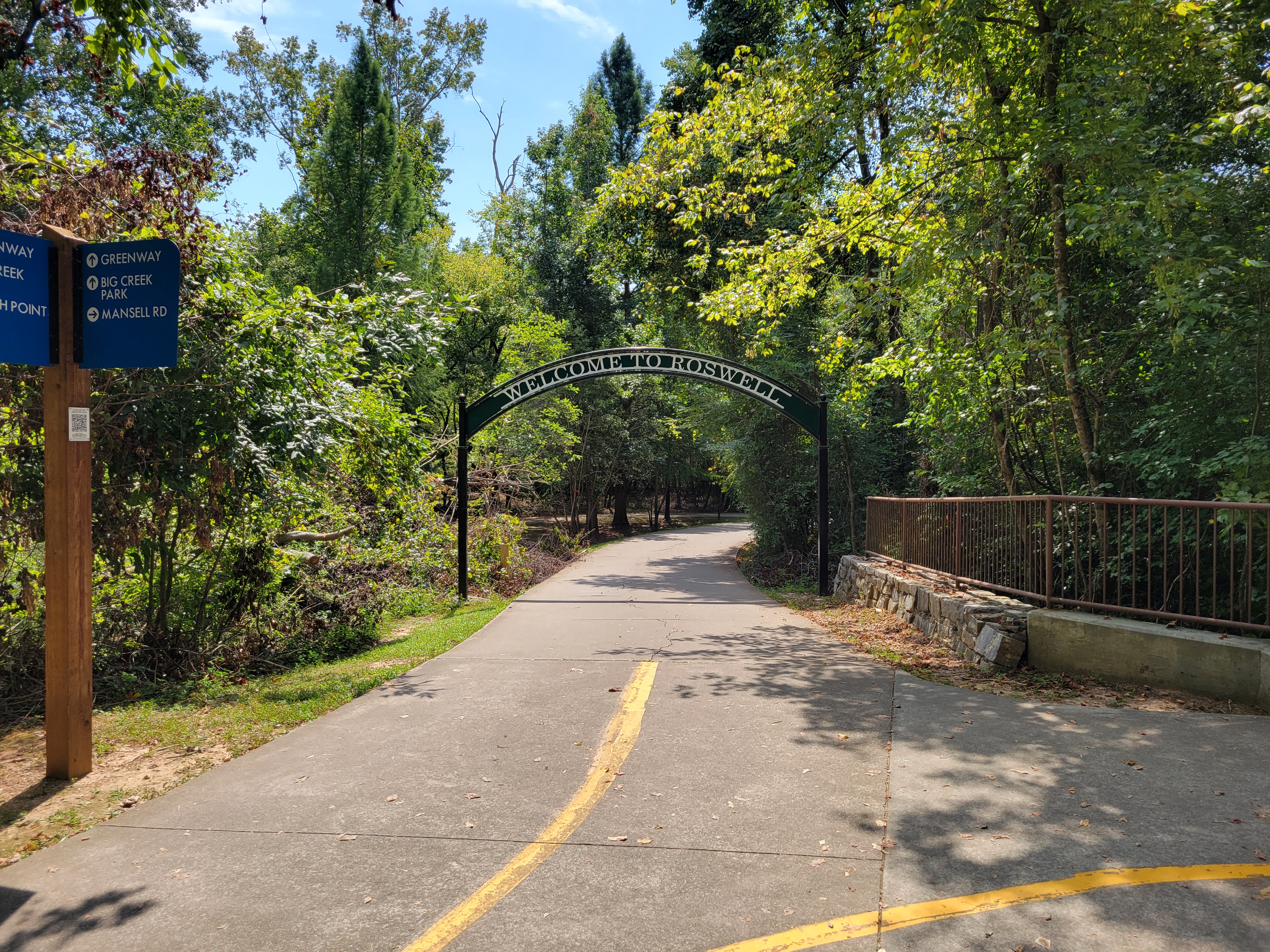 Paved multi-use trail with a Welcome to Roswell arch and directional signage amid trees.