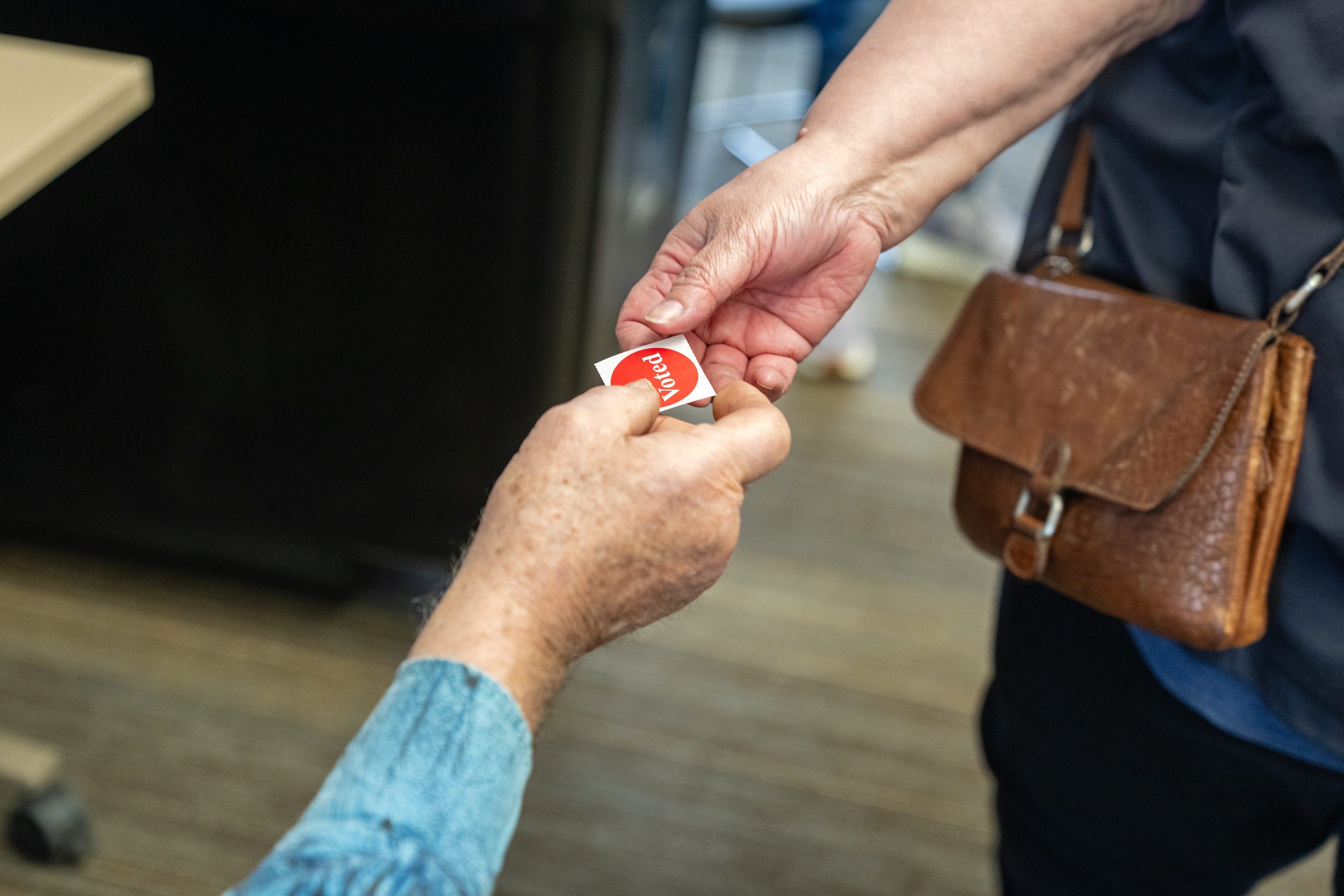 Two hands exchanging a small red 'I Voted' sticker at a polling location.