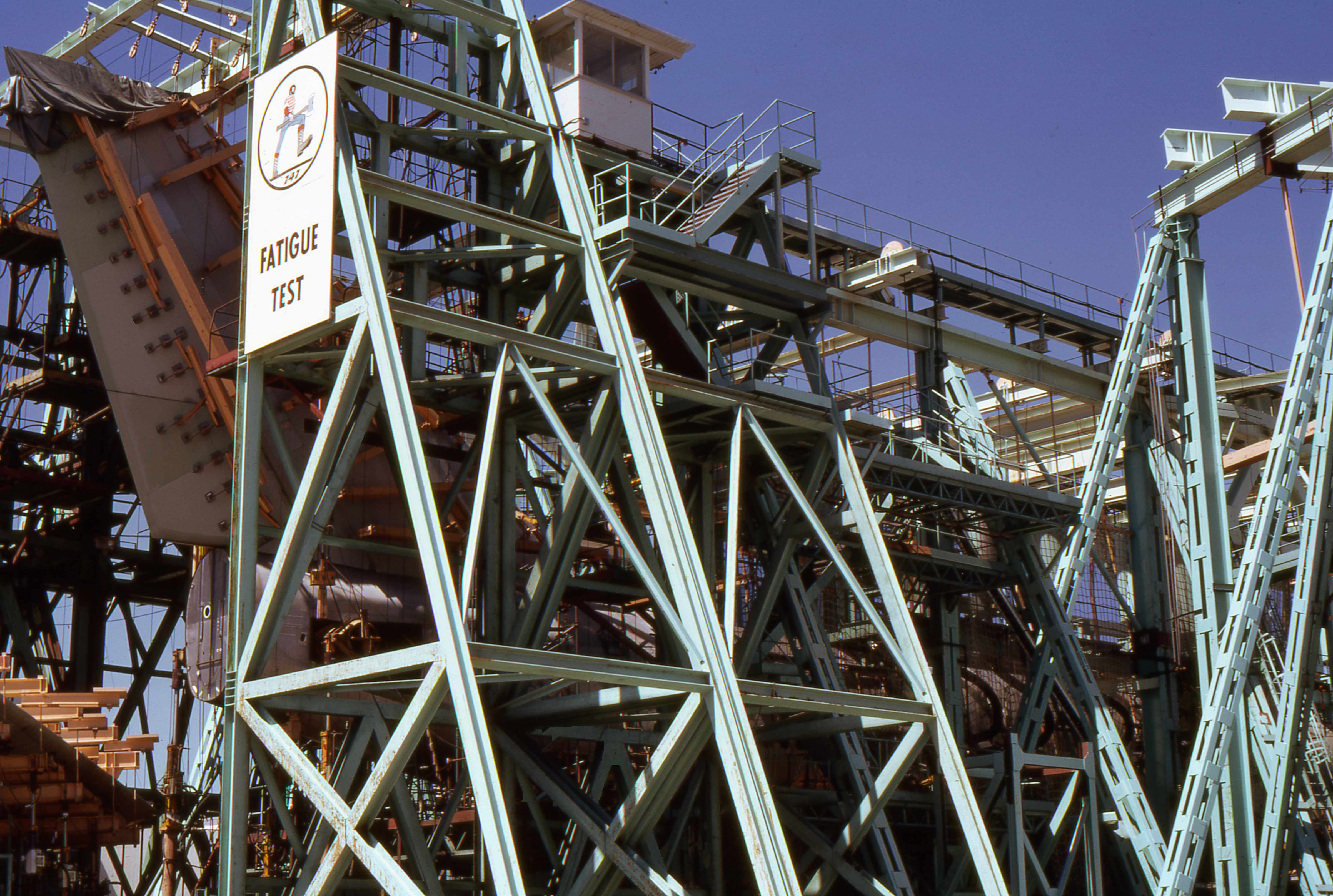 Teal steel truss framework with a fatigue test sign inside a large industrial facility.
