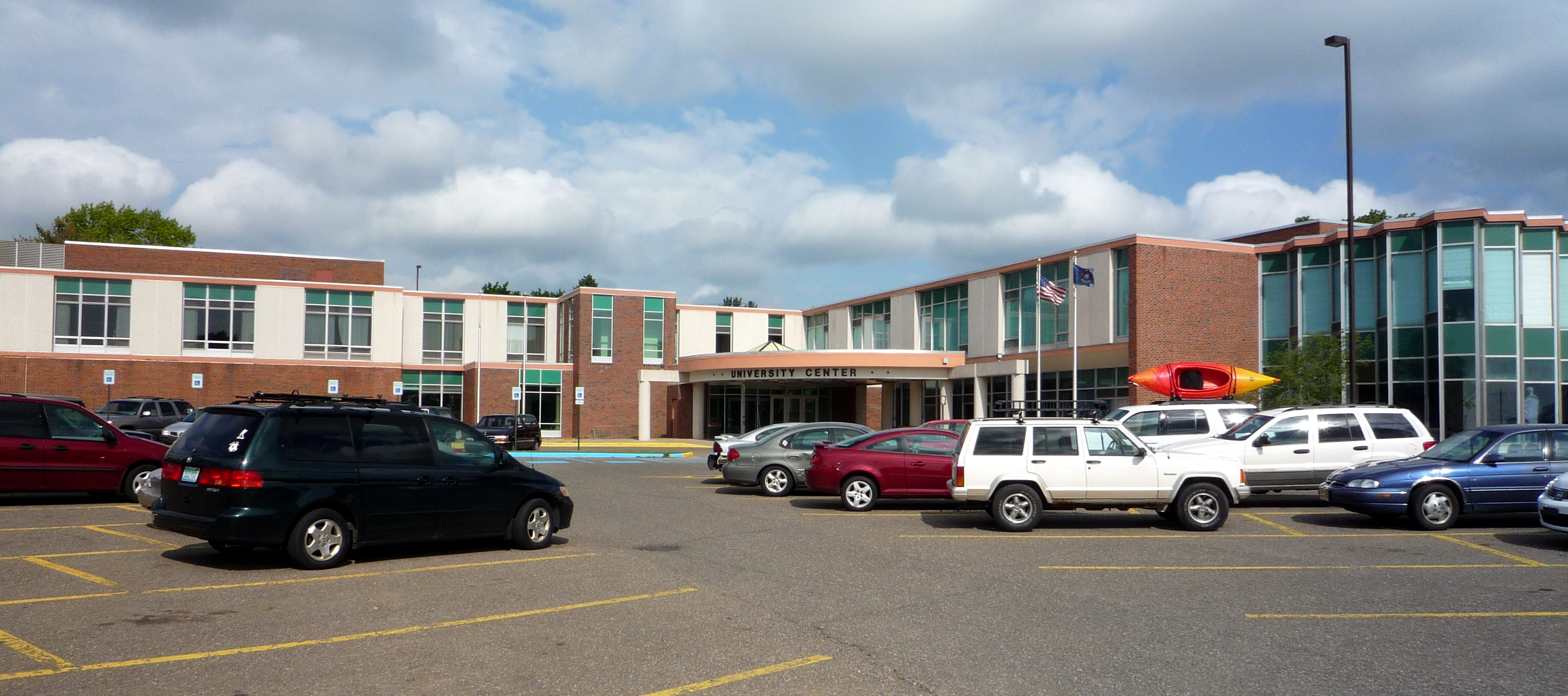 Brick university building with large windows and a parking lot in the foreground.