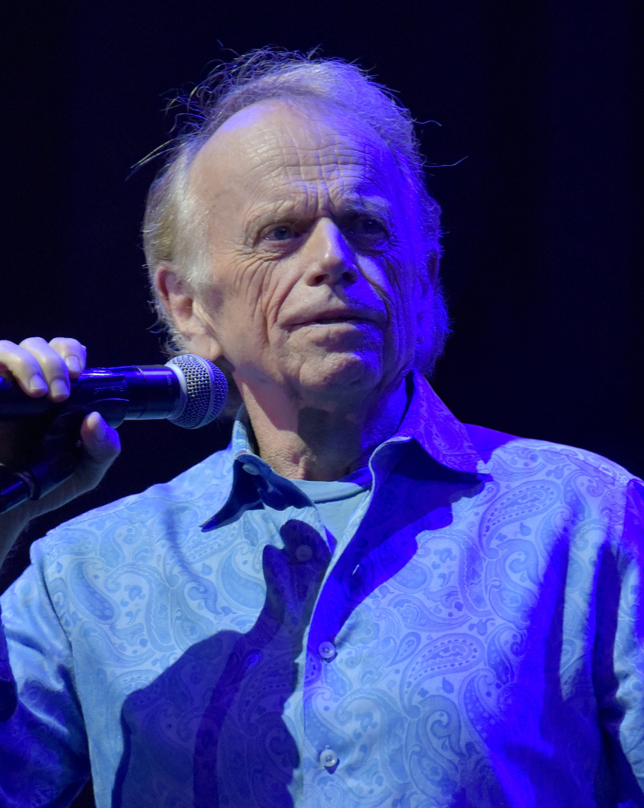 Older man in a paisley shirt holding a microphone under blue-purple stage lighting.