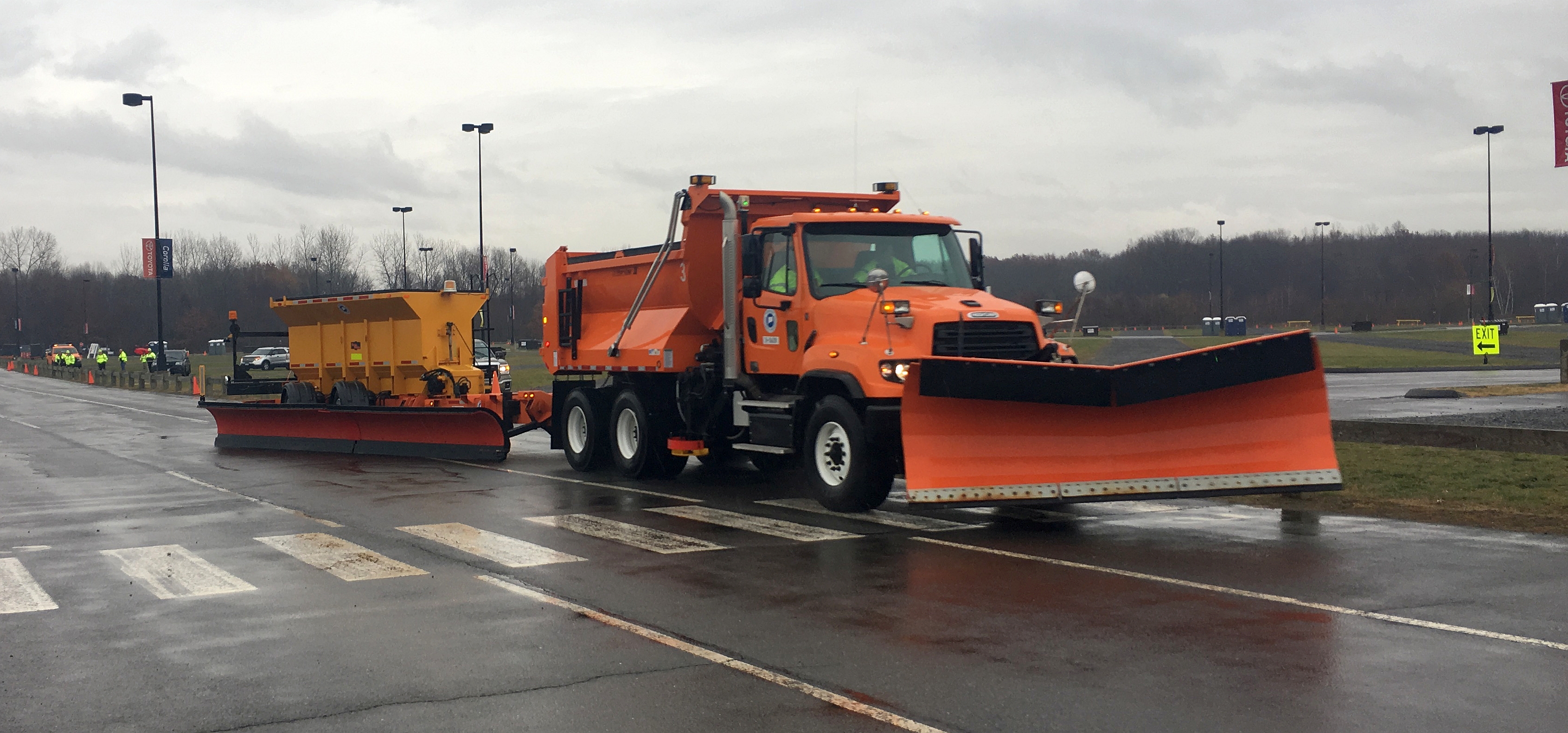 Orange snow plow truck with large front blade and tow-behind plow on wet road.