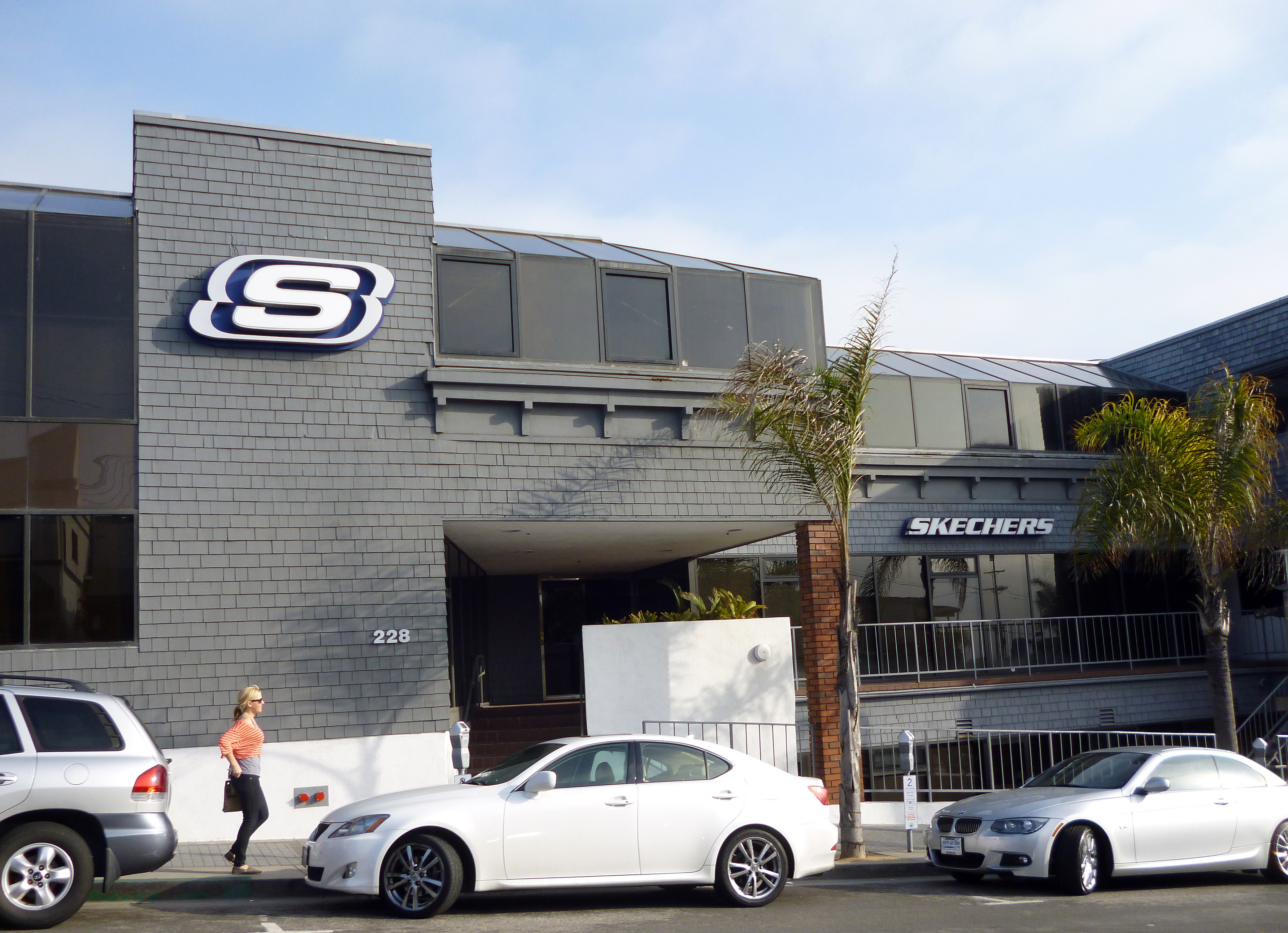 Grey three-story commercial building with a large Skechers logo on the facade and a palm tree out front.