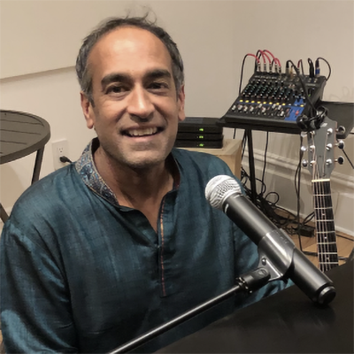 A man smiles at a microphone with an acoustic guitar and mixing board behind him.