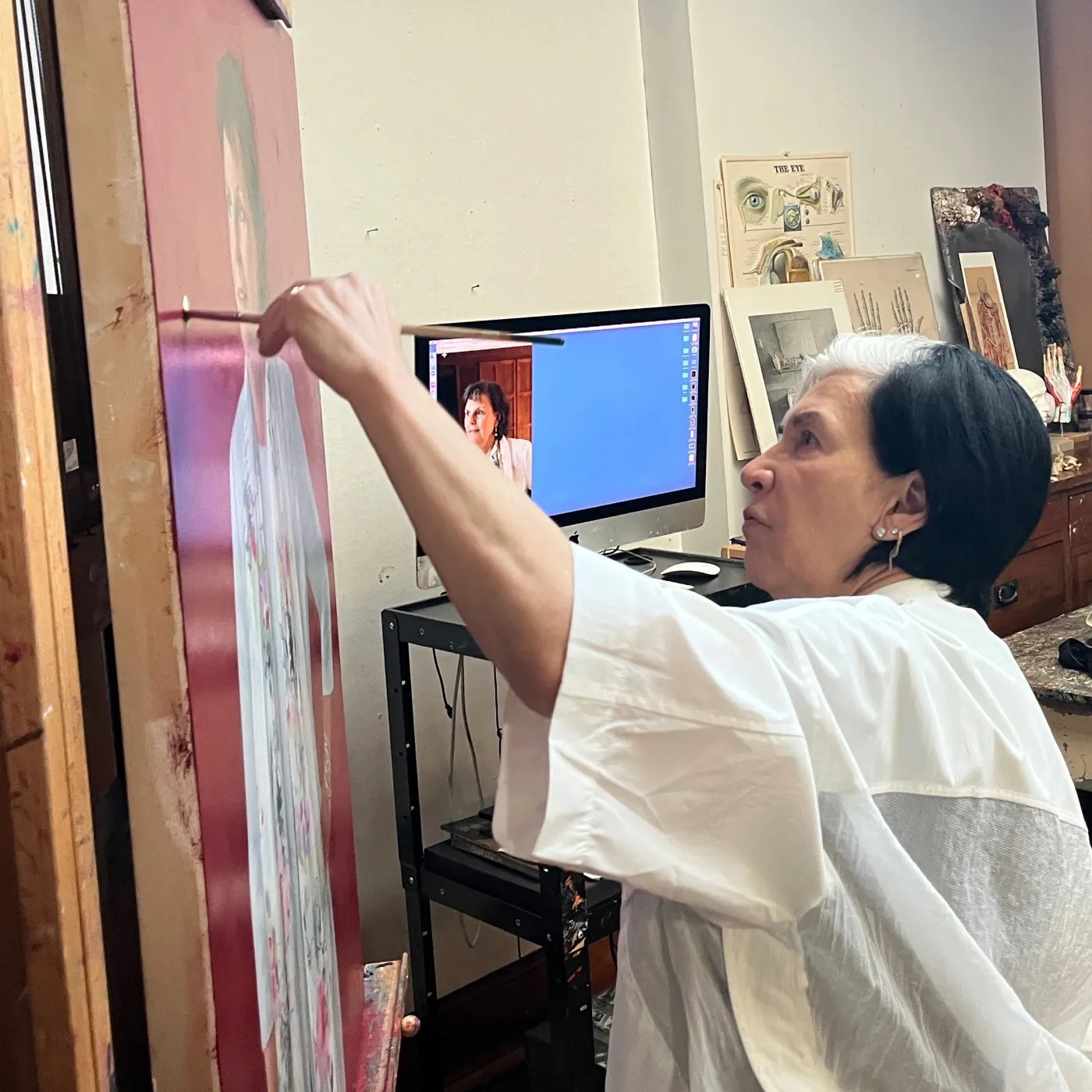 A woman in a white shirt paints a large canvas on a wooden easel in an art studio.