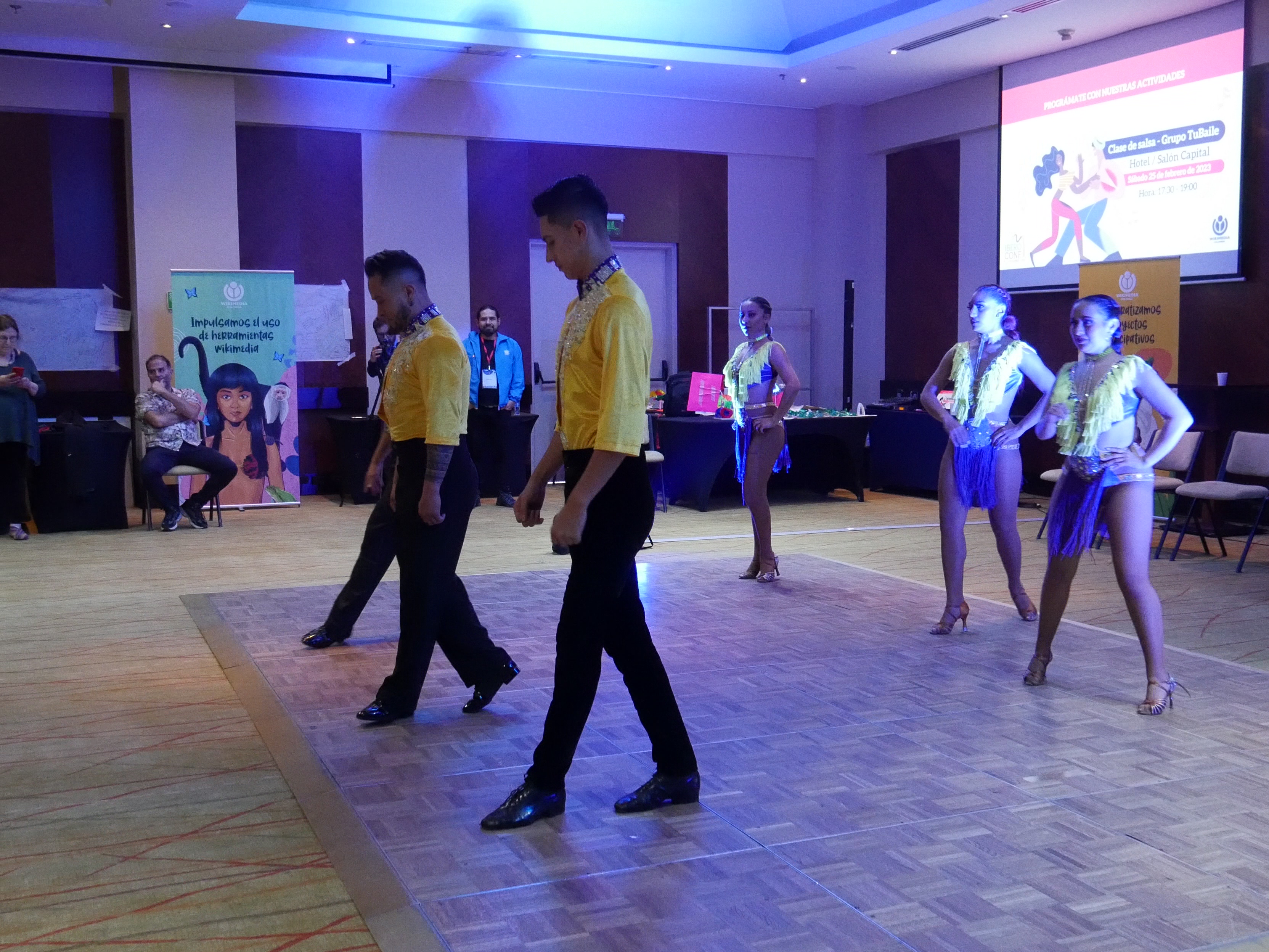 Three male and three female salsa dancers performing in coordinated yellow and blue costumes on a dance floor.