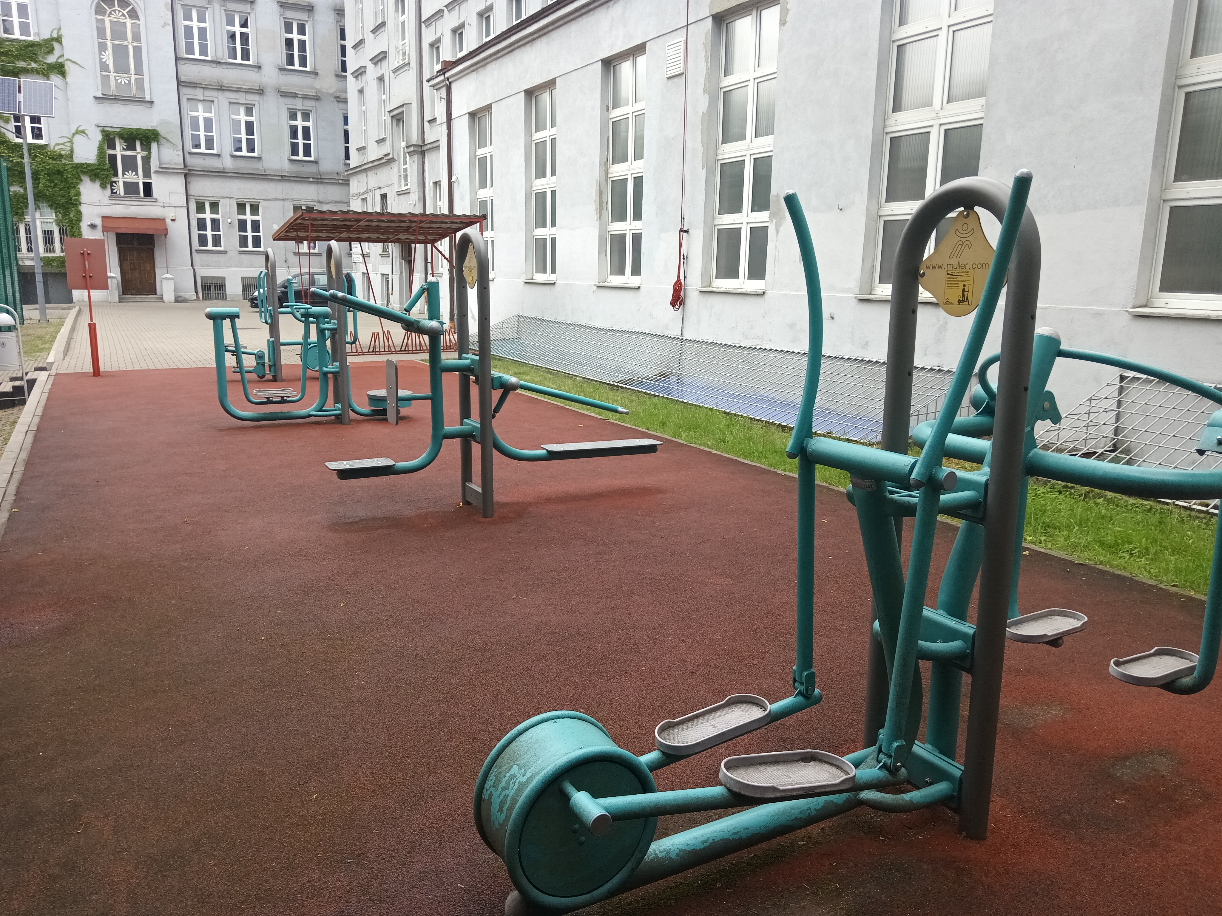 Teal outdoor fitness equipment including an elliptical machine on a red rubberized surface.