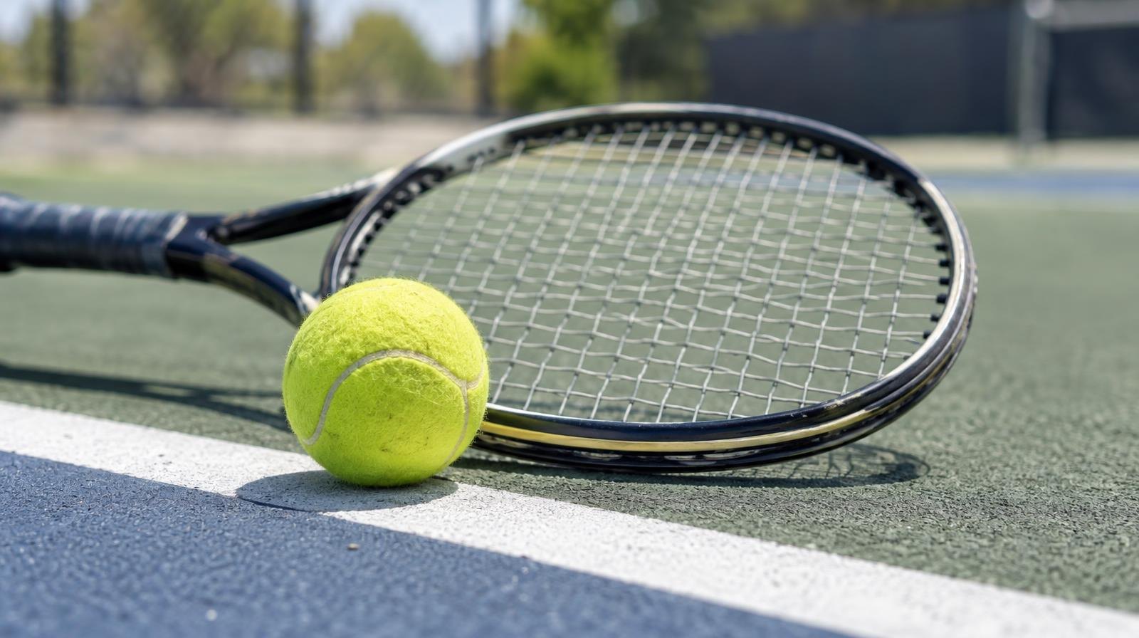 A tennis racket and yellow tennis ball resting on a hard court surface near the baseline.