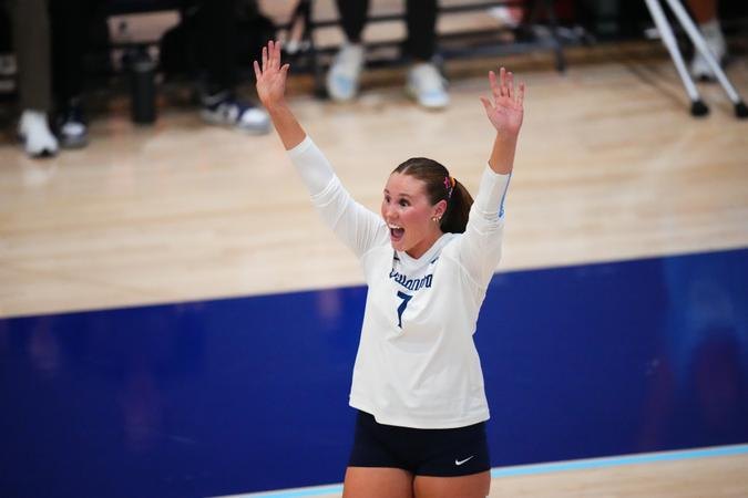 Volleyball player in white jersey raises both arms in celebration on a blue indoor court.