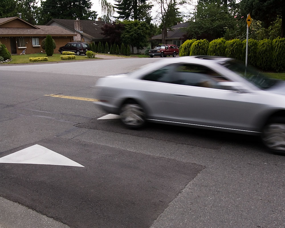 A silver sedan drives over raised asphalt speed cushions at a residential intersection.