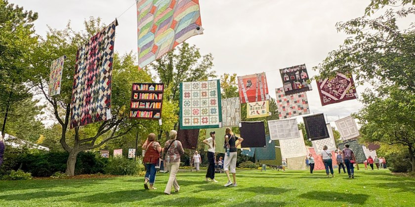Dozens of colorful patchwork quilts hanging on lines outdoors among green trees.