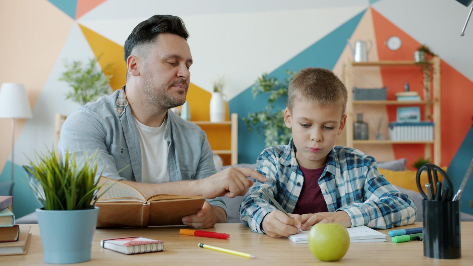 Father guiding his son during homework session in a colorful room.