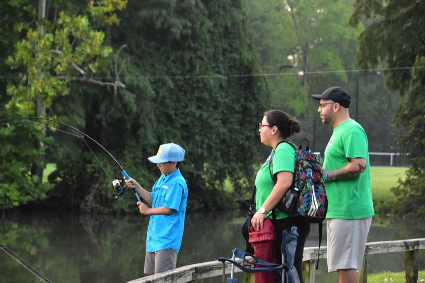 A child in a blue shirt casts a fishing rod near a pond while two adults watch.