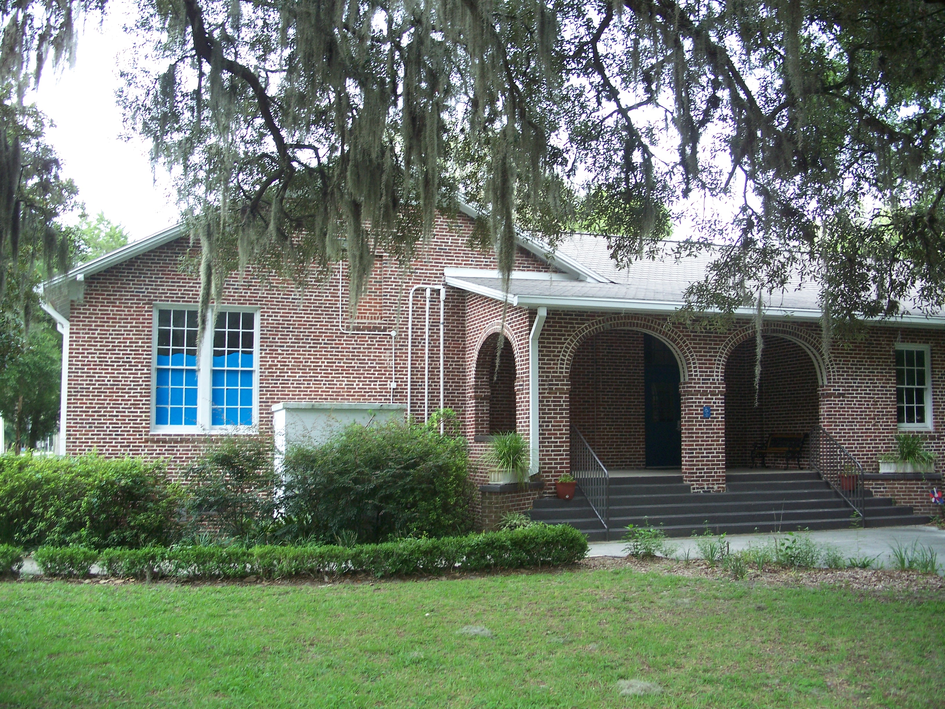 Single-story red brick school building with arched columns and Spanish moss-draped trees.