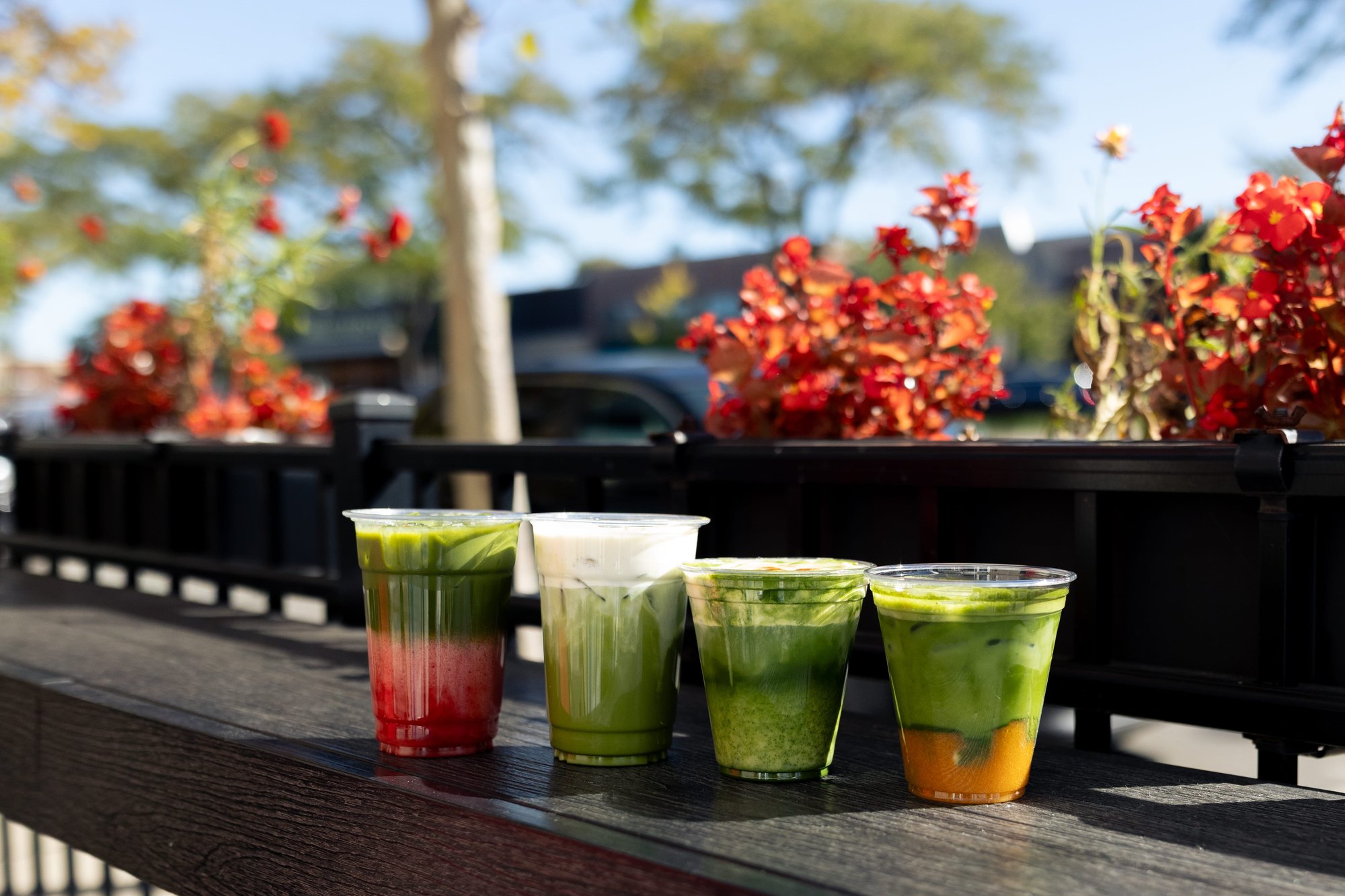 Four layered green and red matcha drinks in clear cups on an outdoor railing with flowering plants behind.