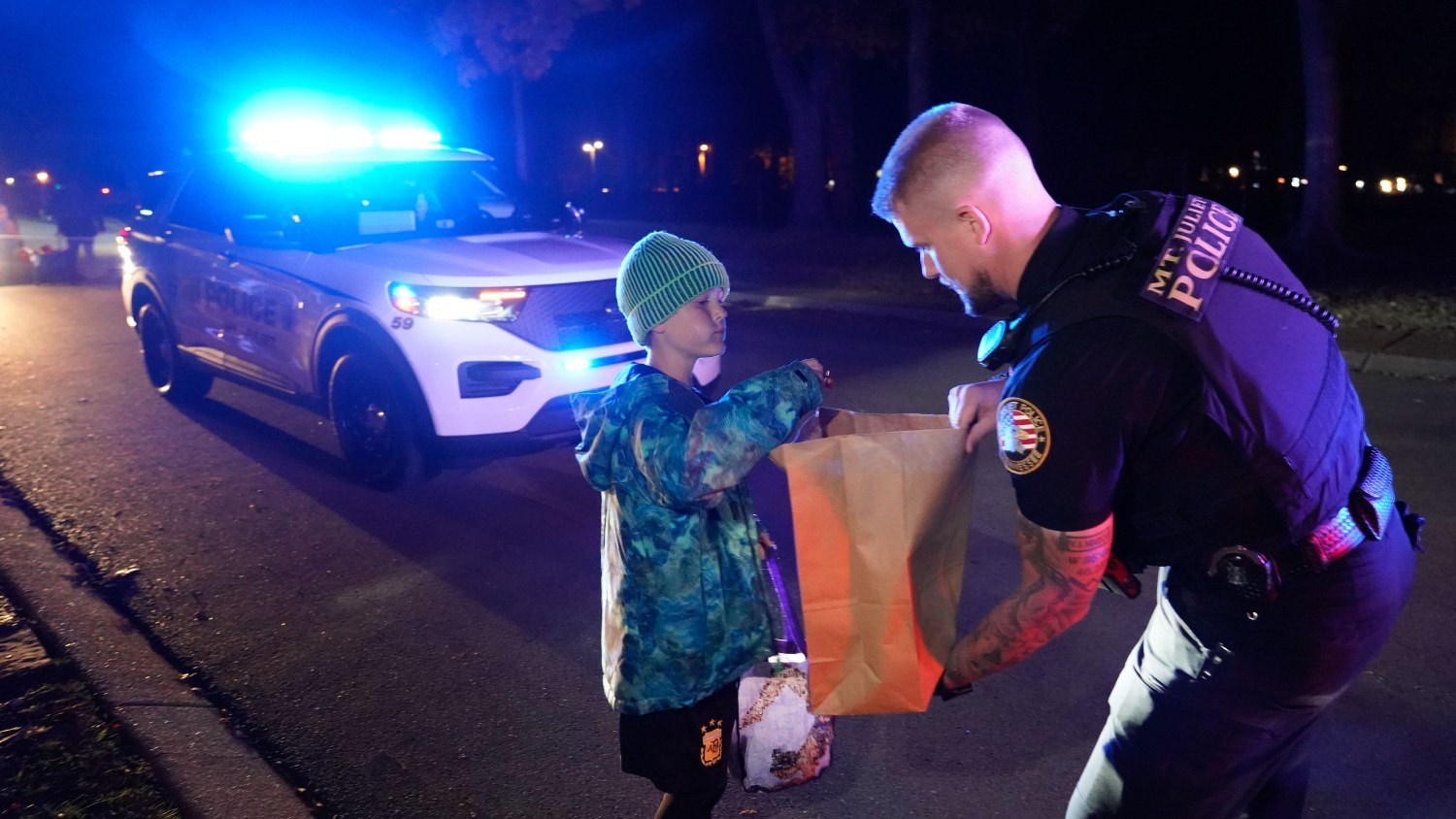 A uniformed officer hands a paper bag to a child at night near a police SUV with blue lights on.