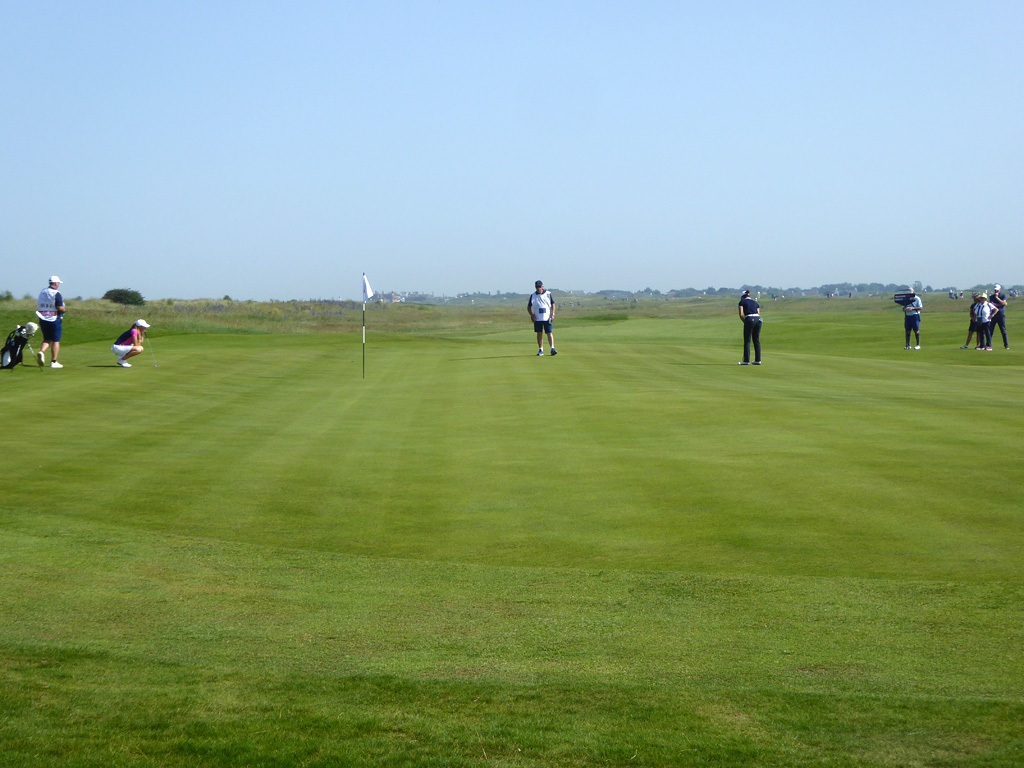 Several golfers and caddies on a golf course green under a clear blue sky.
