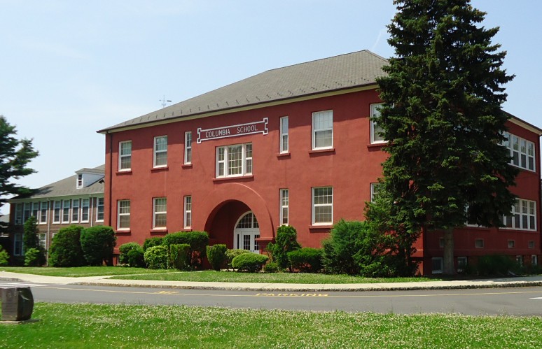 Two-story red brick school building with arched entrance and evergreen trees.