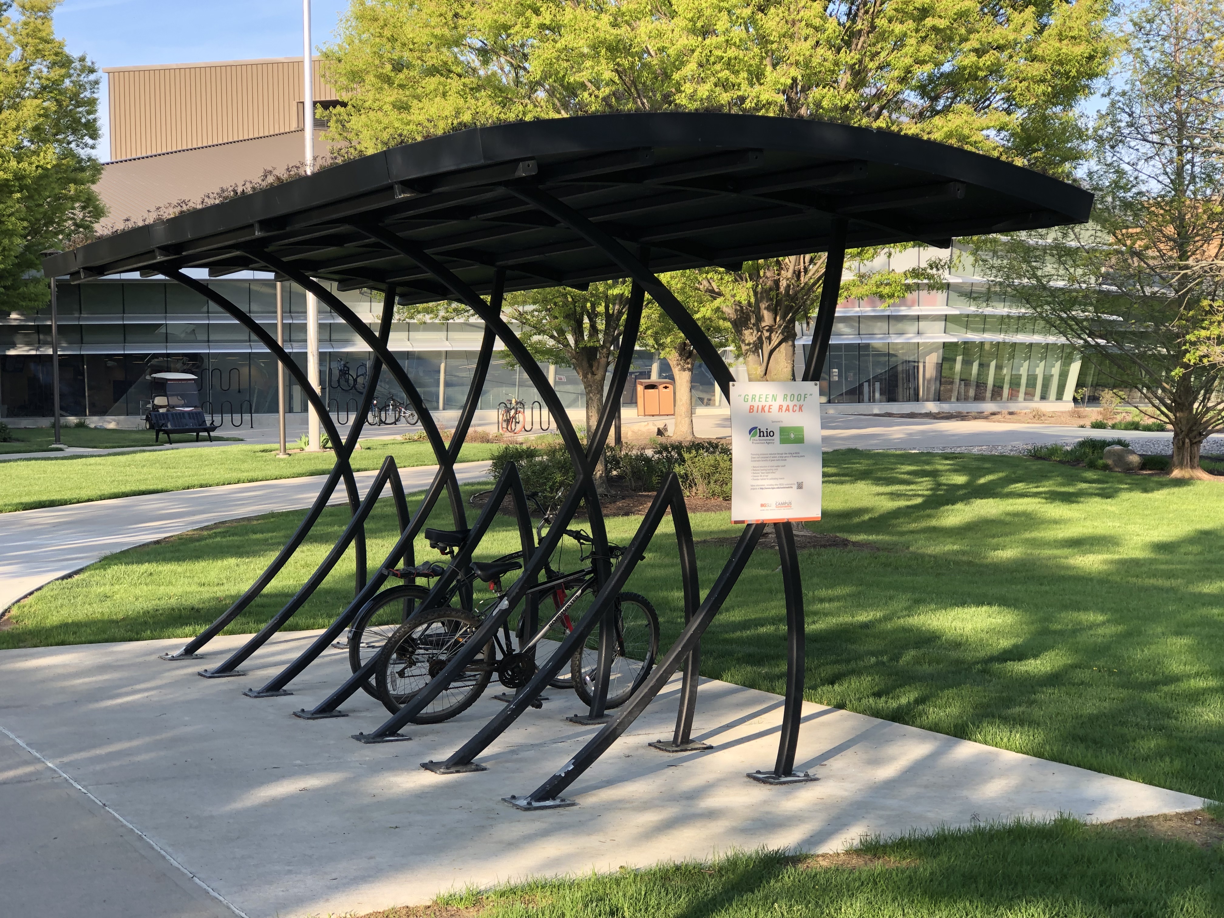 Covered bicycle shelter with a curved green roof beside a grassy campus lawn.
