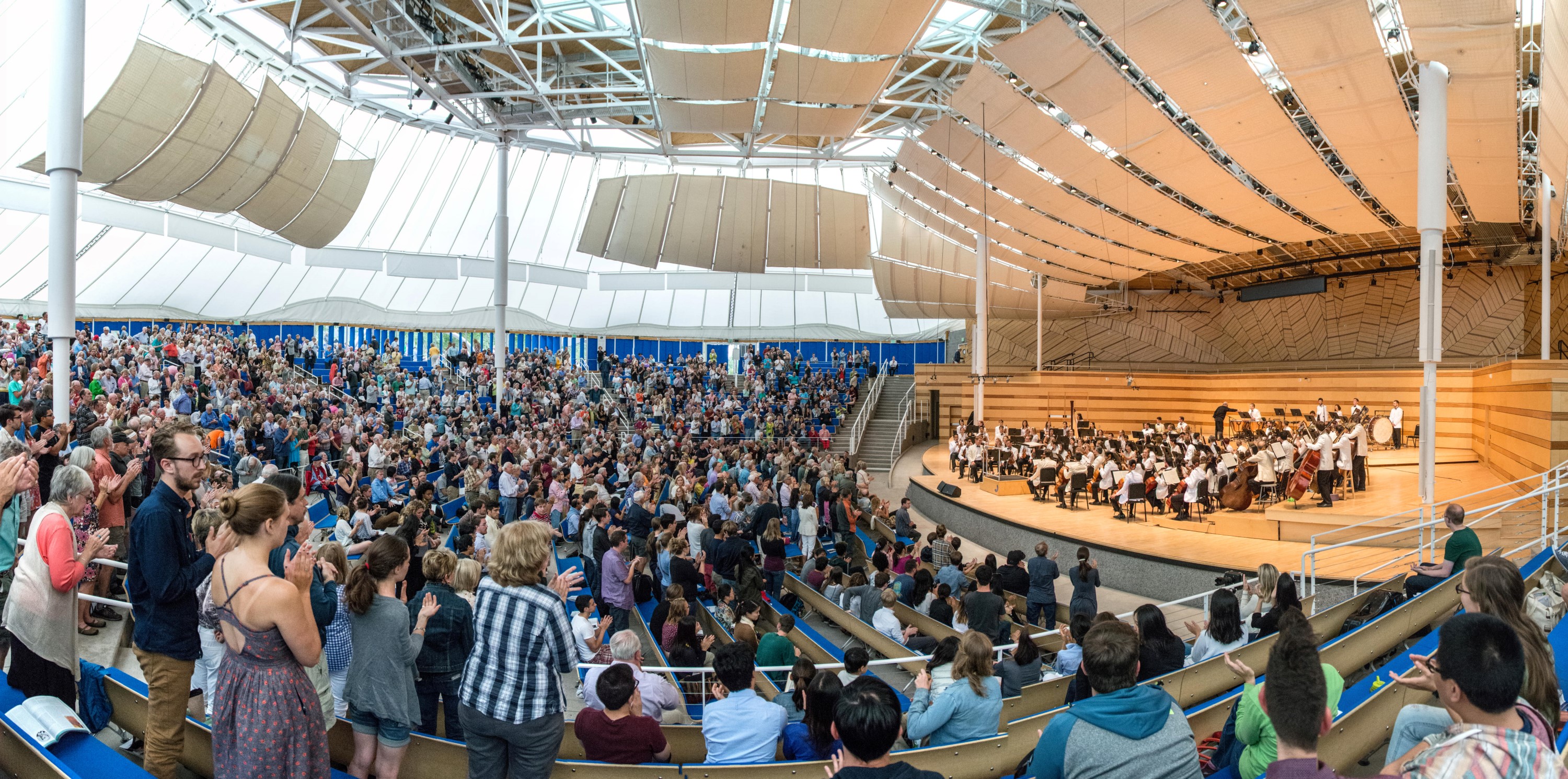 Hundreds of audience members fill an outdoor amphitheater as an orchestra performs on stage.